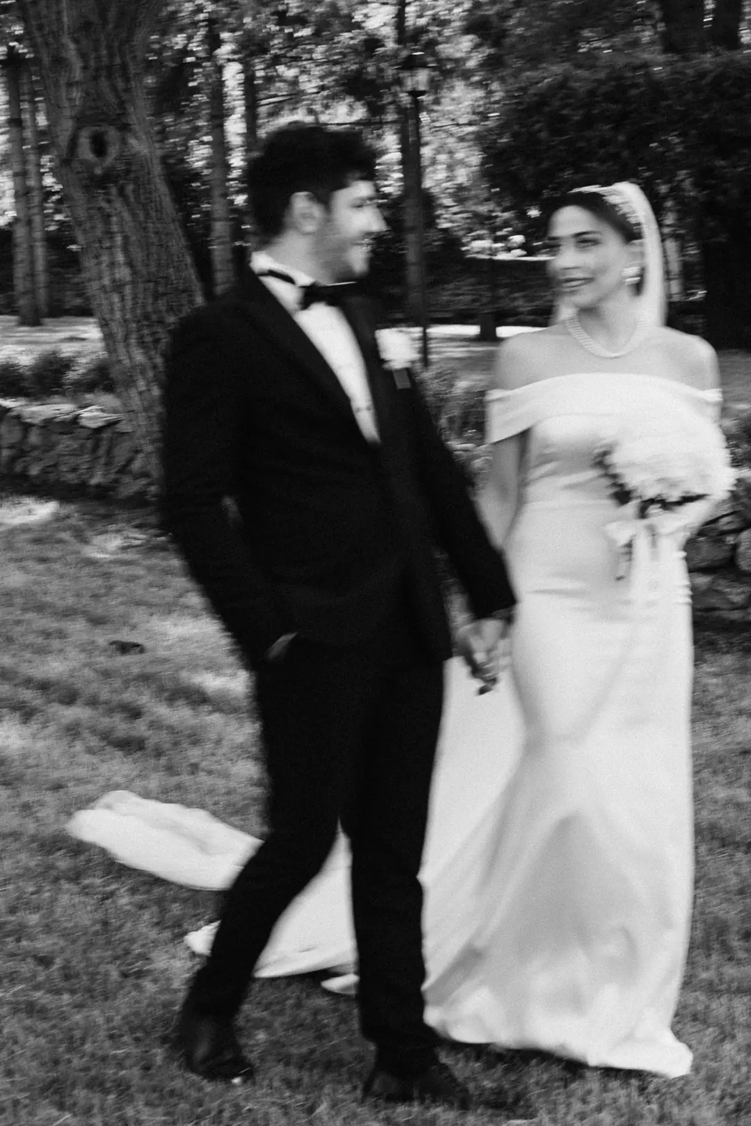 Musa and Solmaz walking hand in hand across a lawn under tall trees, in black and white