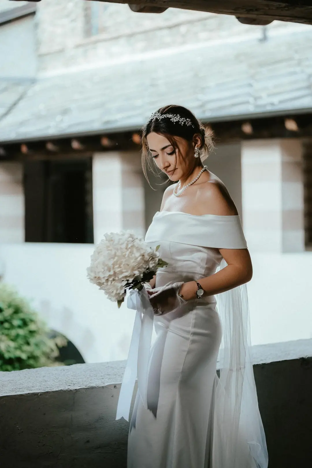 Solmaz con un abito di raso a spalle scoperte e una collana di perle guarda un bouquet di ortensie bianche sotto un portico di legno