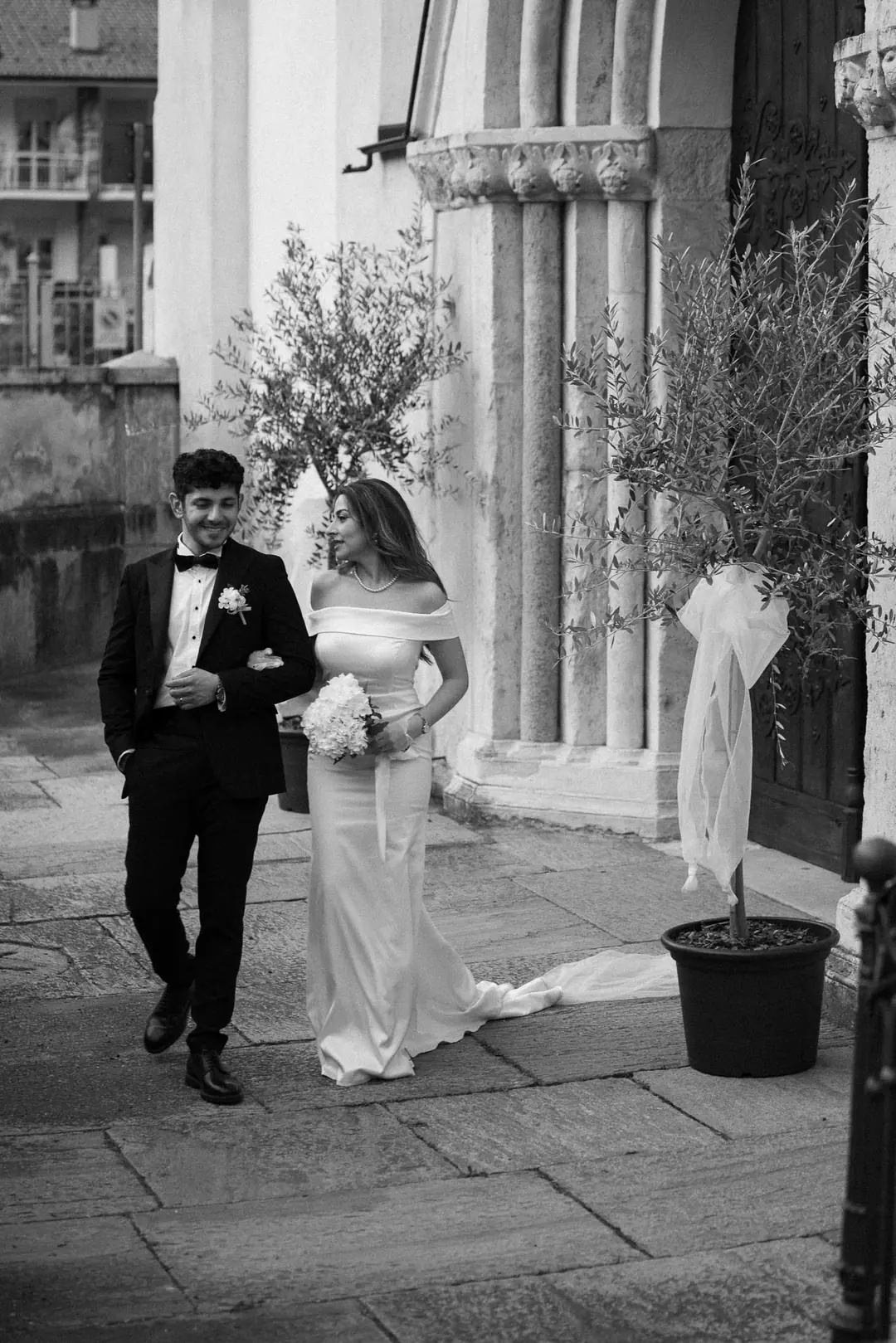 Musa and Solmaz walking arm in arm past a carved stone church doorway and potted olive trees tied with white ribbon, in black and white