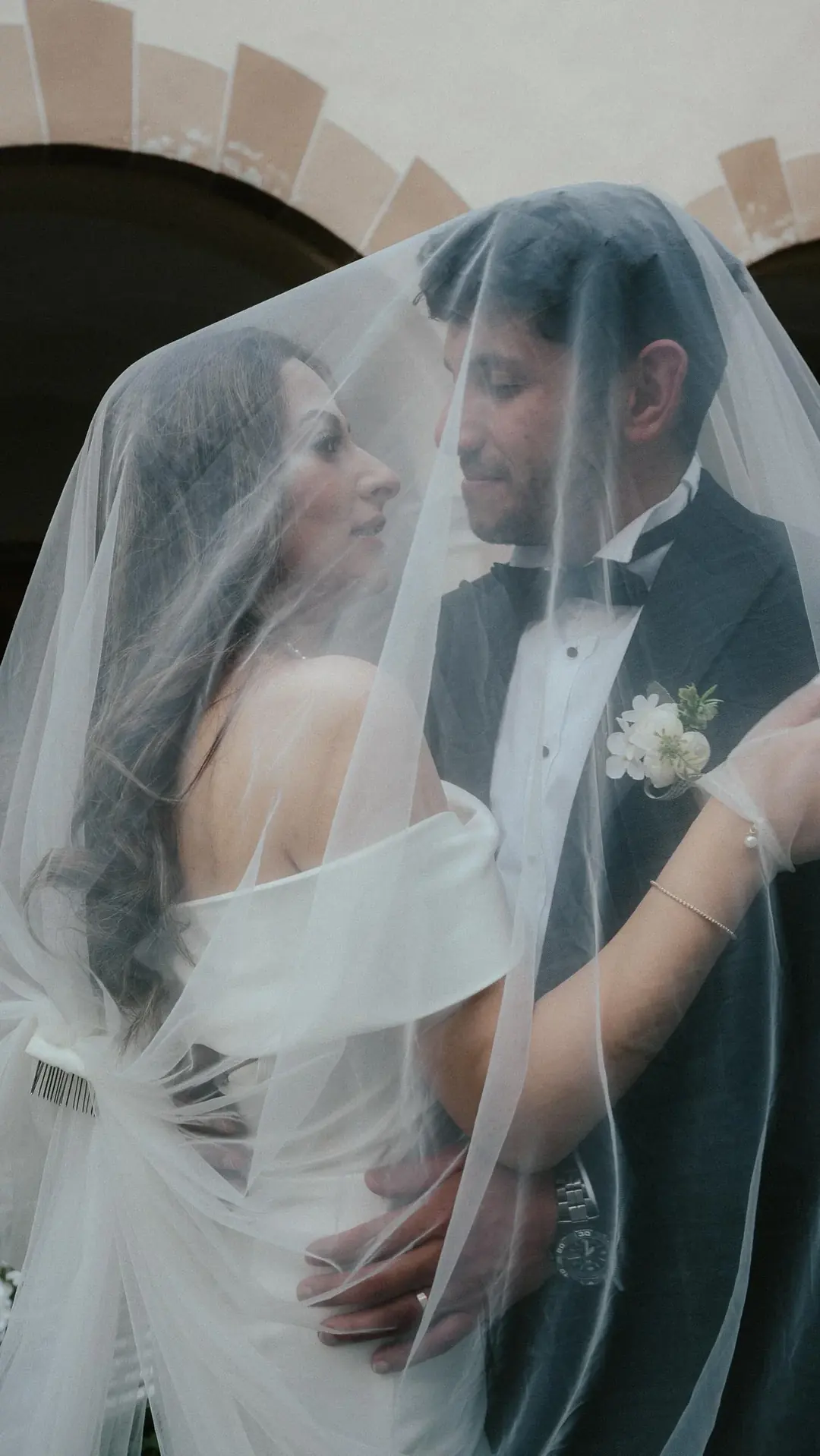 A couple close together under her veil beneath a stone arch edged in pale brick, in Susa