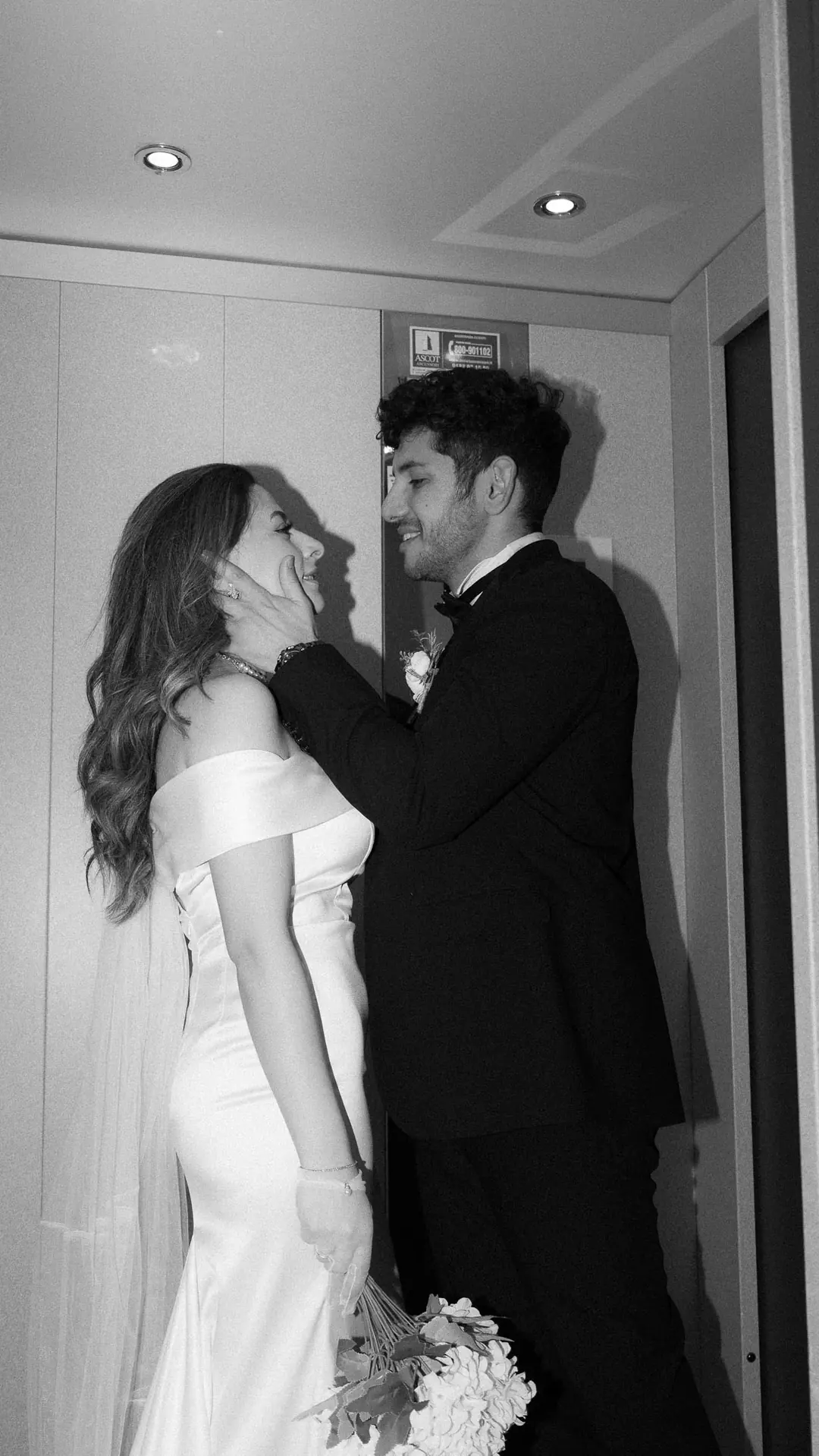 Musa holding Solmaz's face as she laughs with her eyes closed in a hotel lift, white hydrangeas in her hand, in black and white