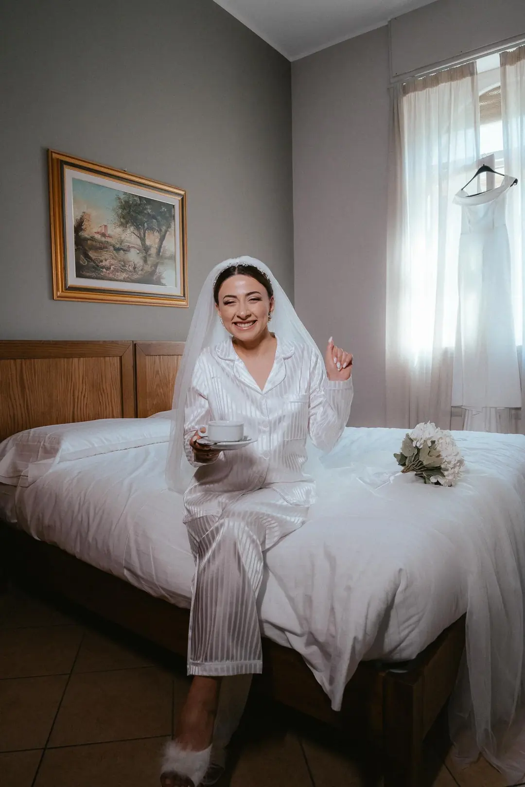 Solmaz smiling on the edge of a hotel bed in striped pyjamas and a veil, holding a cup of coffee, a white gown hanging in the window