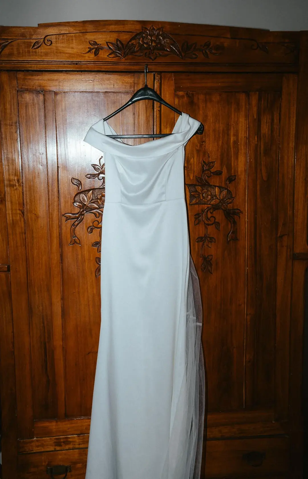 A white off-the-shoulder wedding gown hanging on the carved door of a wooden wardrobe