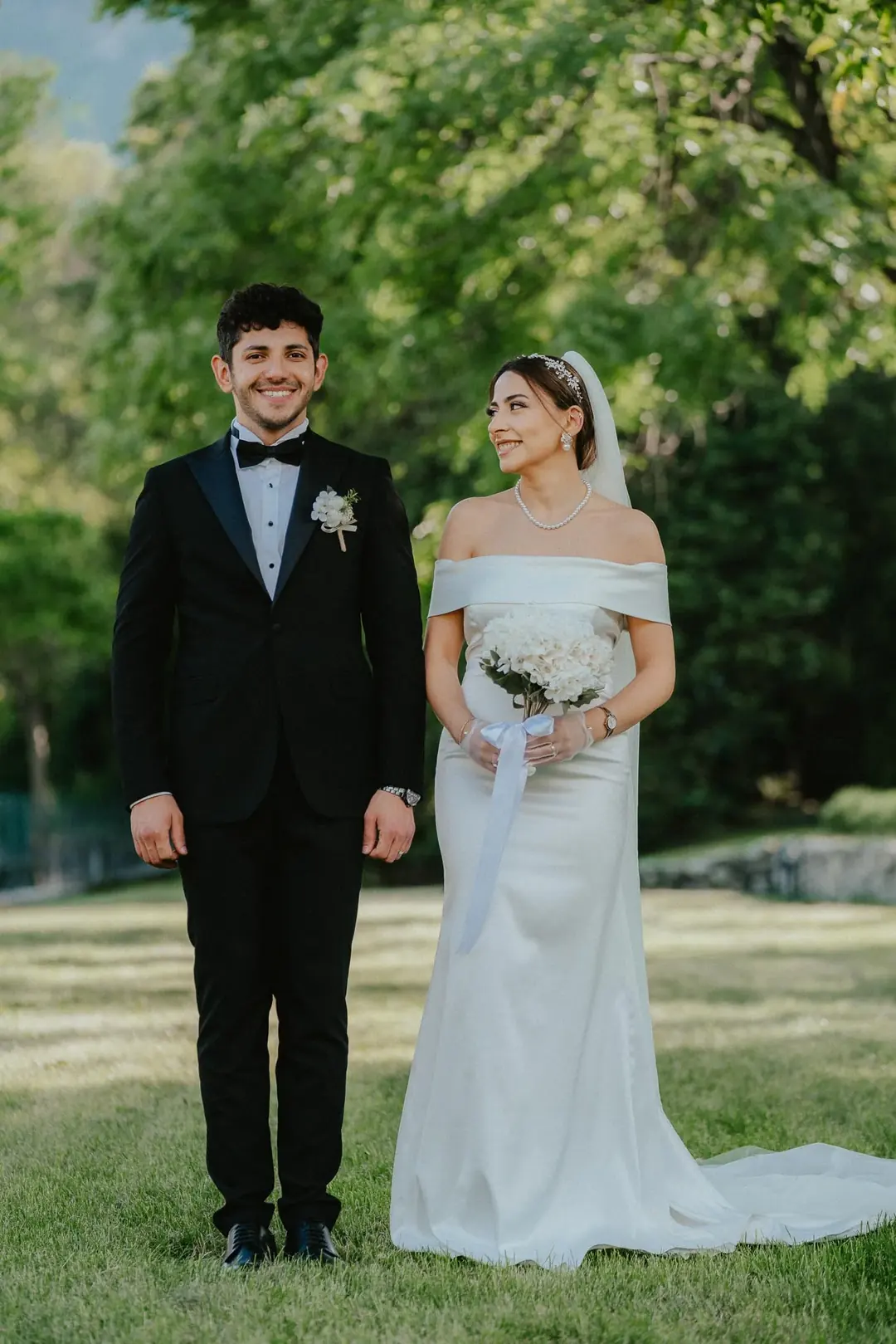 Musa and Solmaz standing side by side on a lawn under tall trees in Susa, Piedmont, she in an off-the-shoulder gown holding white hydrangeas
