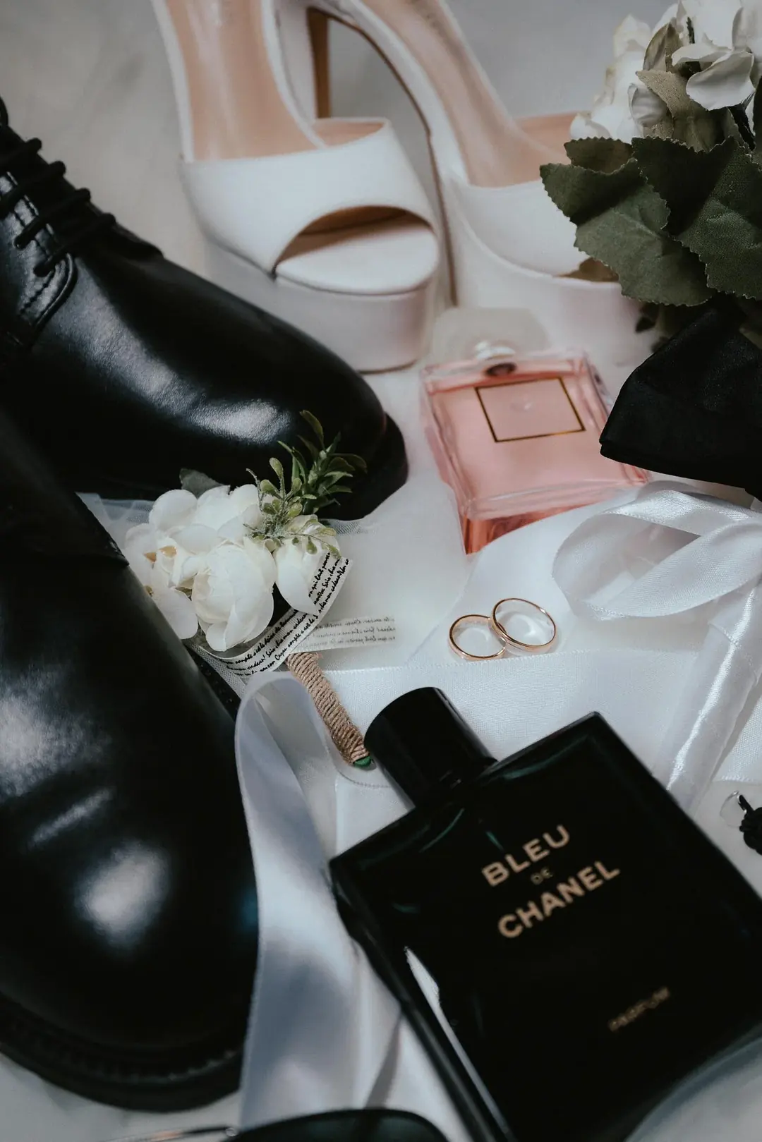 Two gold wedding rings on white fabric beside a bride's white heels, a groom's black shoes, a white boutonniere and two perfume bottles, photographed in Susa