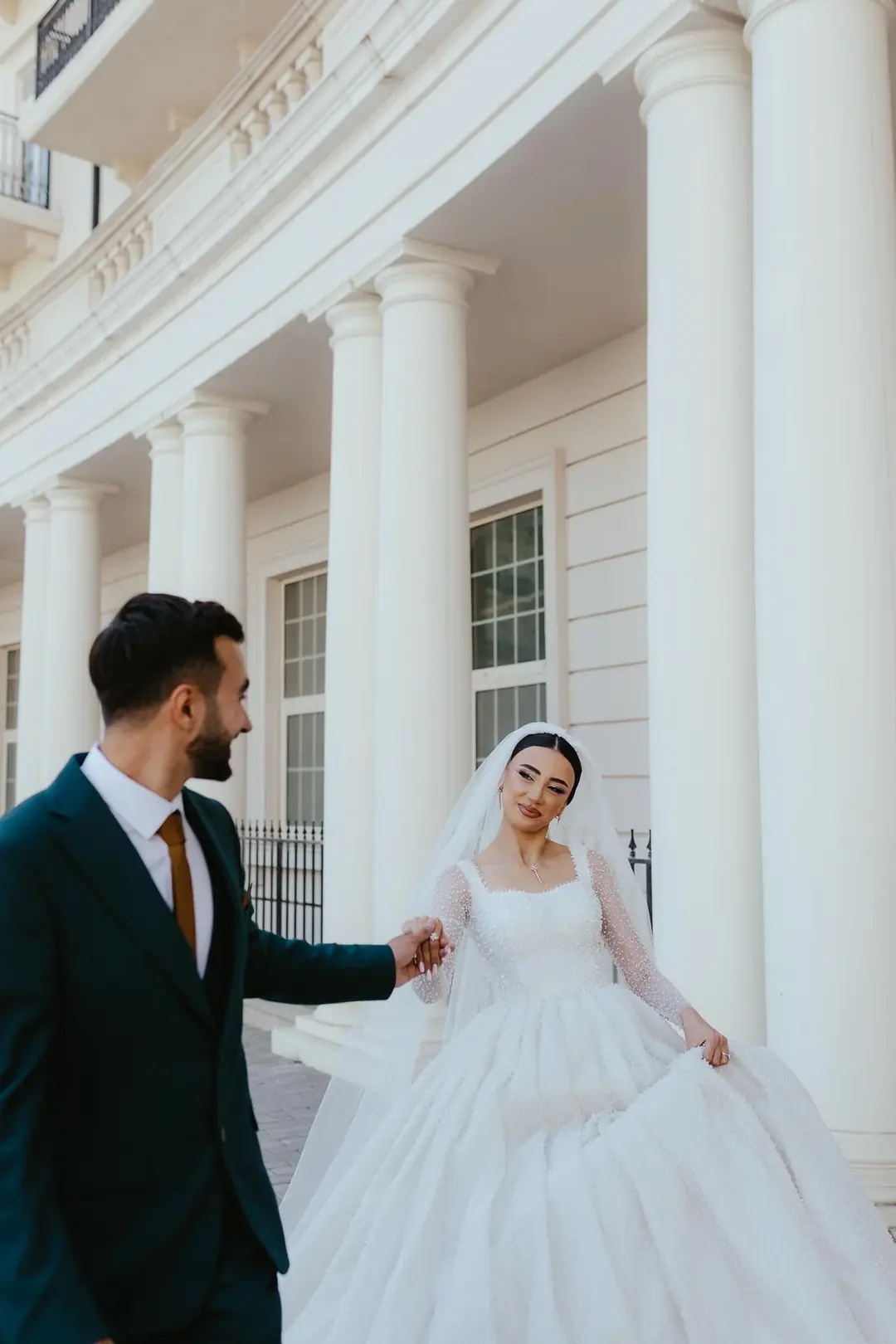 A groom leading the bride by the hand past tall white columns, her full pearl-covered gown gathered in her other hand