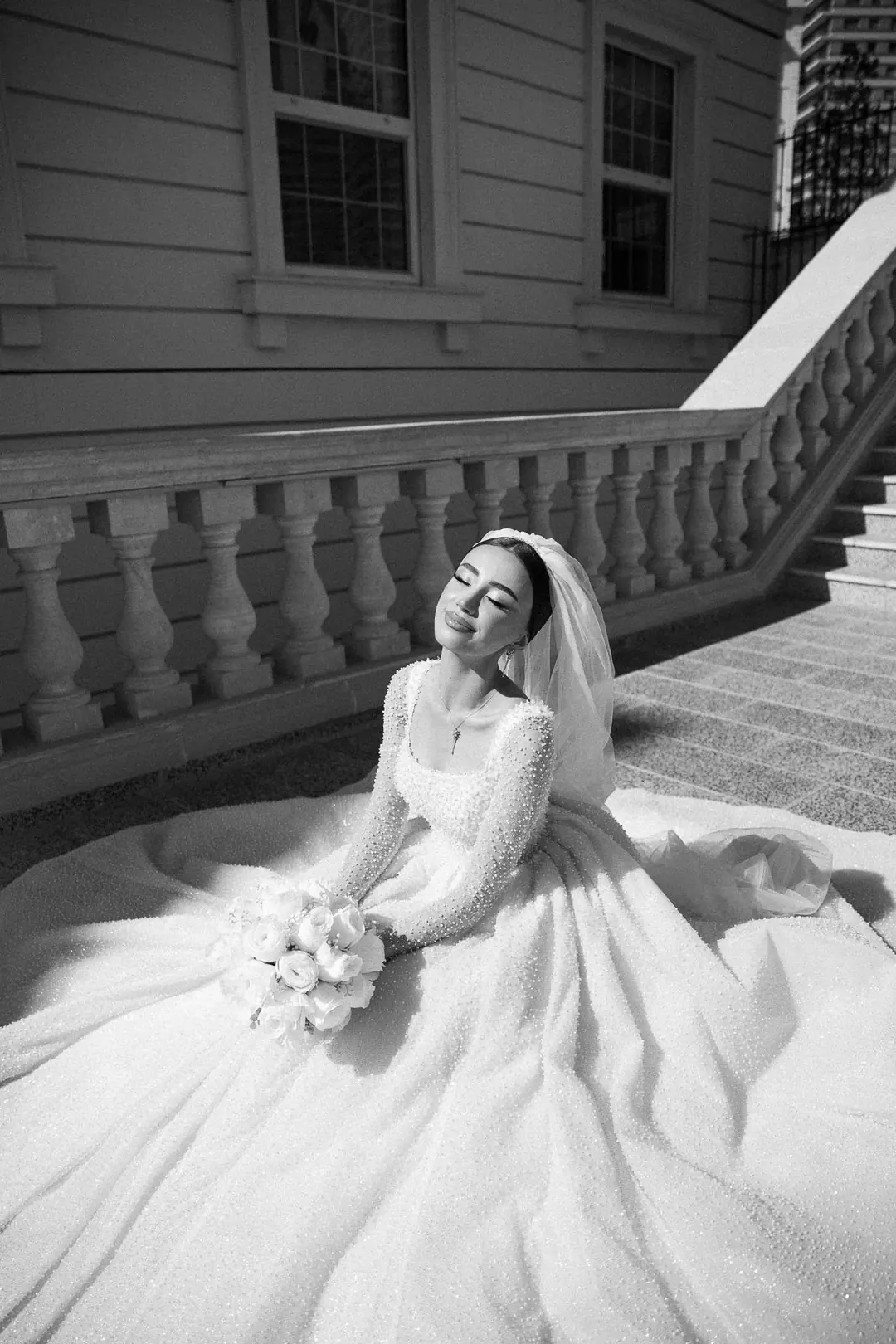 Narmin sitting on a paved terrace by a stone balustrade, her full pearl-covered gown spread around her, eyes closed and smiling in the sun, in black and white