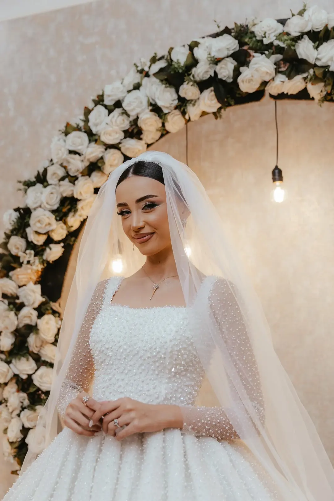 Narmin smiling in a pearl-covered gown and long veil, standing in front of an arch of white roses with bare bulbs hanging beside her, Baku