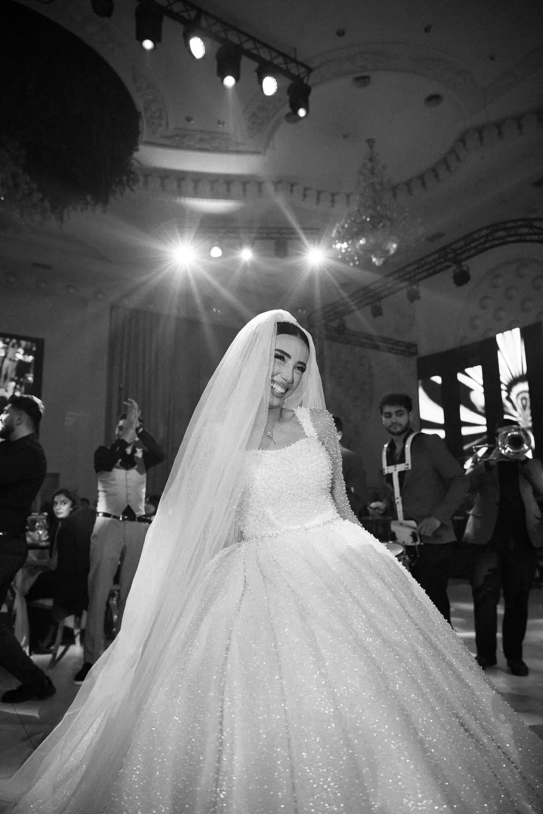 A bride laughing on the dance floor in her pearl-covered gown, musicians, one with a trumpet, playing behind her under stage lights, black and white