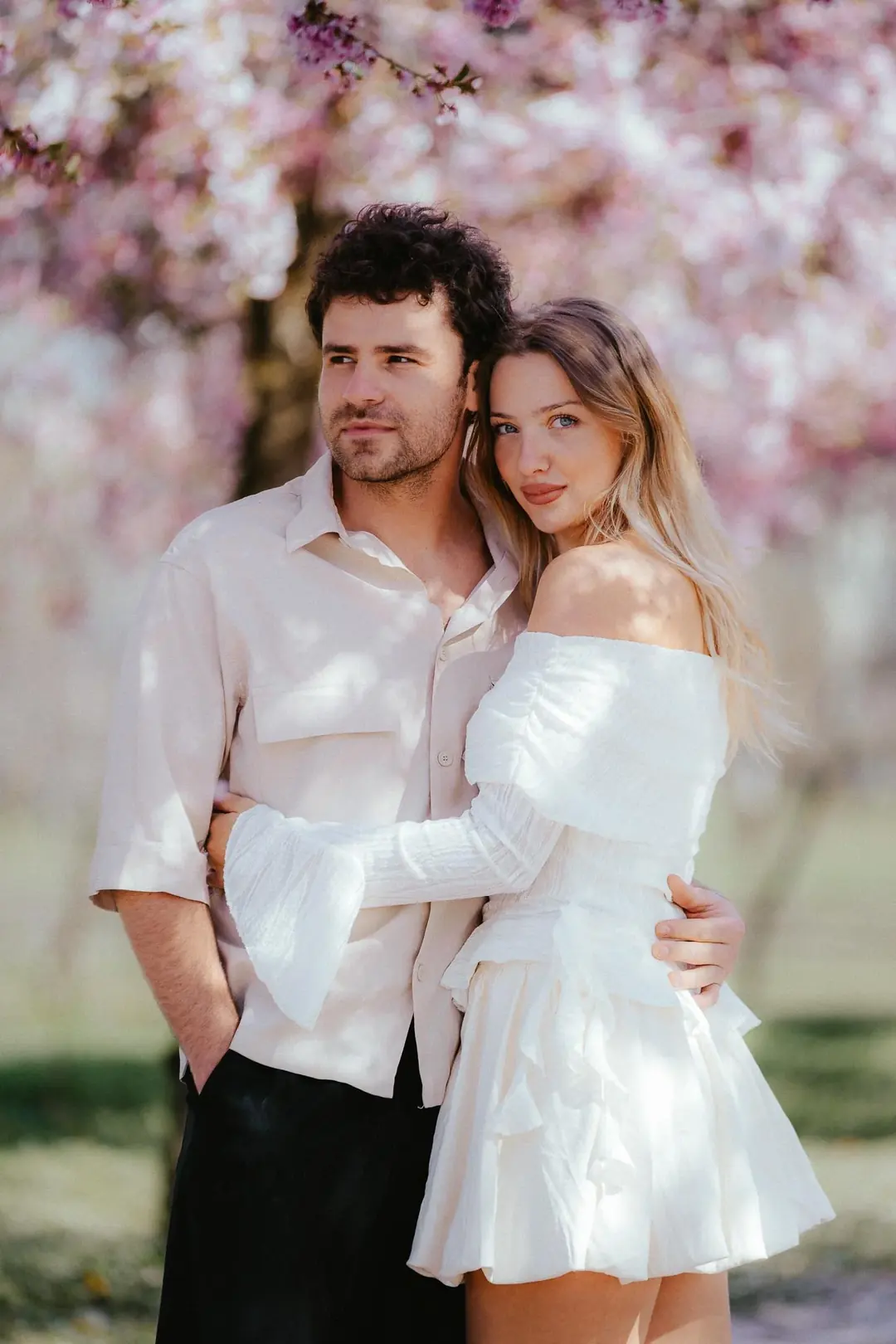 Gabri and Mada standing close under cherry trees in full pink blossom in Turin, she in a white ruffled dress