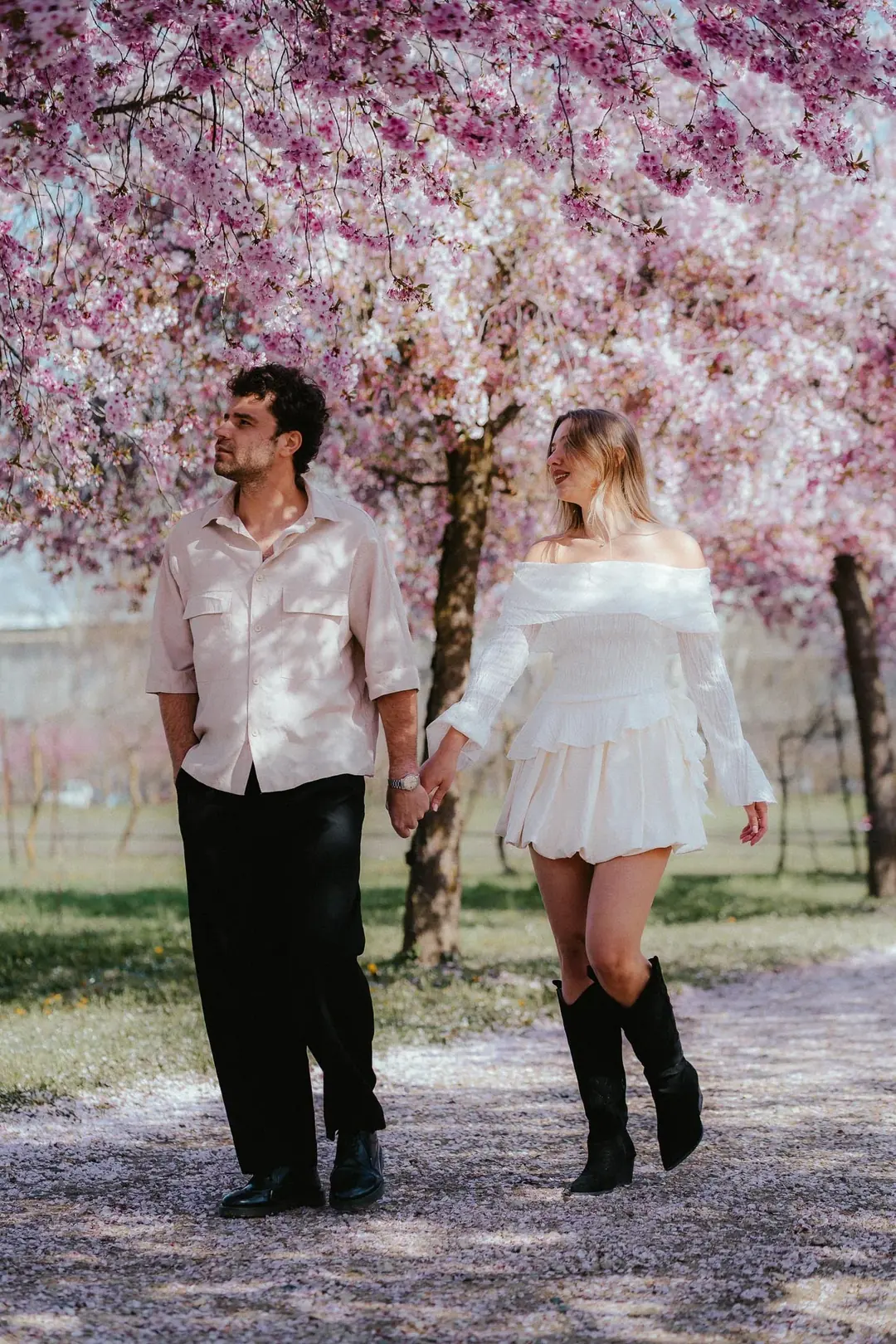 Gabriele e Maddalena camminano mano nella mano sotto i ciliegi in fiore rosa in un parco di Torino, con i petali sul sentiero