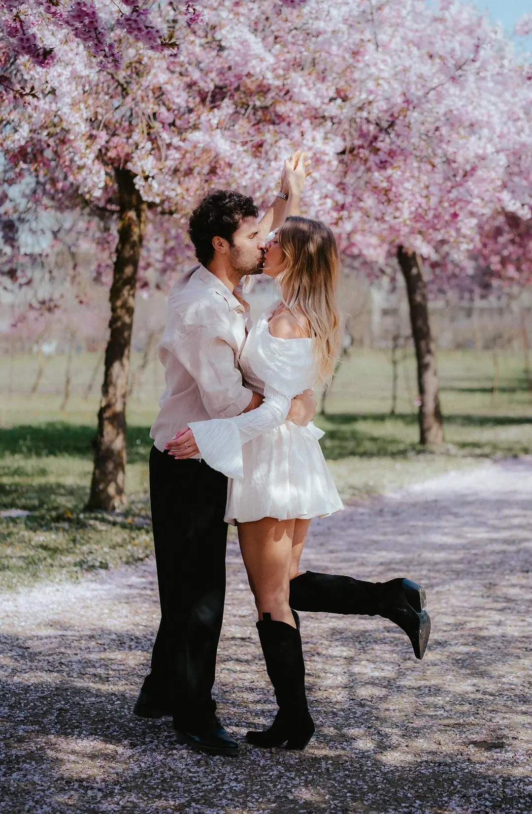 Gabri and Mada kissing on a path covered in fallen cherry petals, her boot lifted, pink blossom overhead