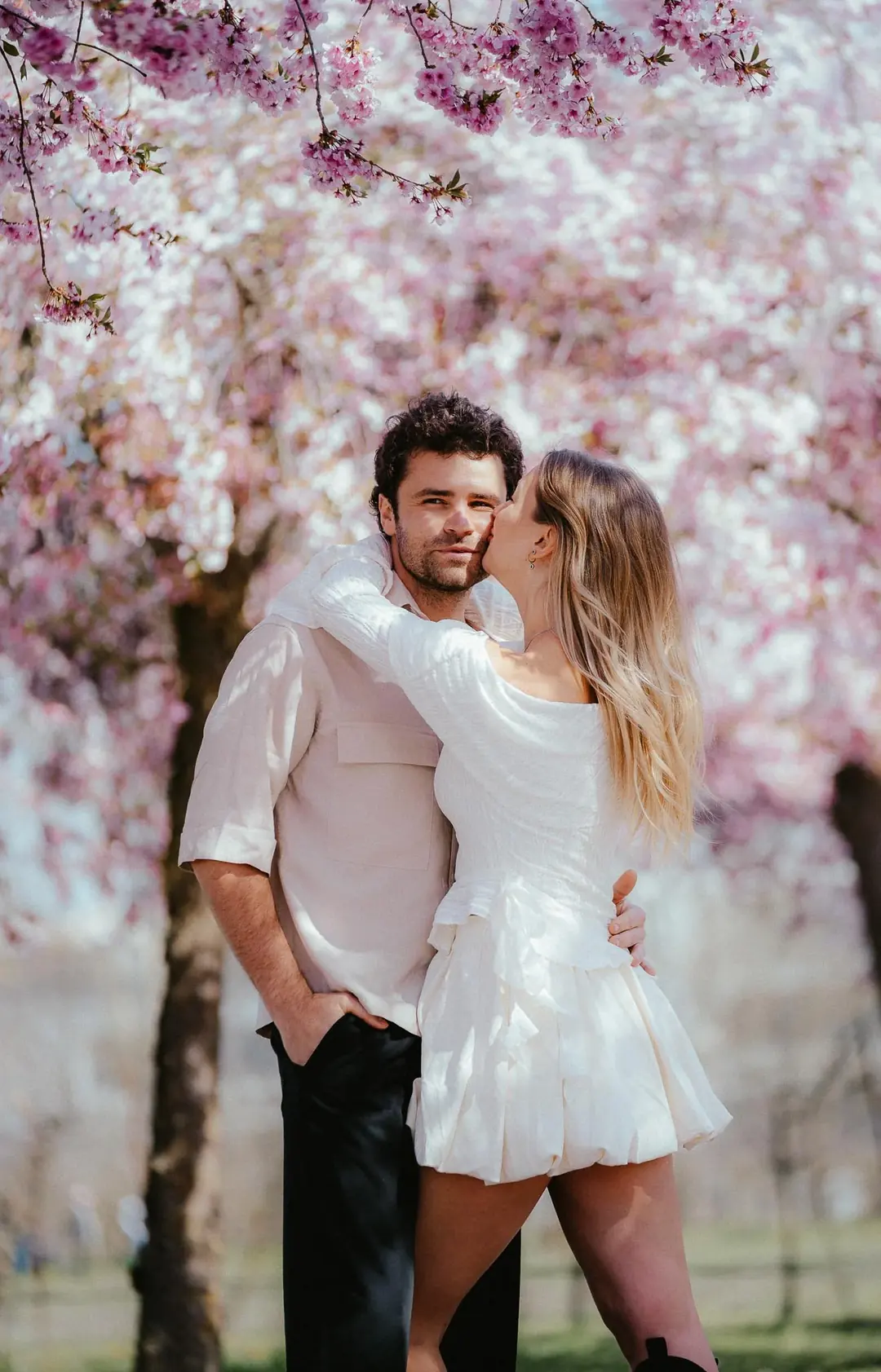 Gabri and Mada under pink cherry blossom in Turin, Mada kissing his cheek as he looks at the camera