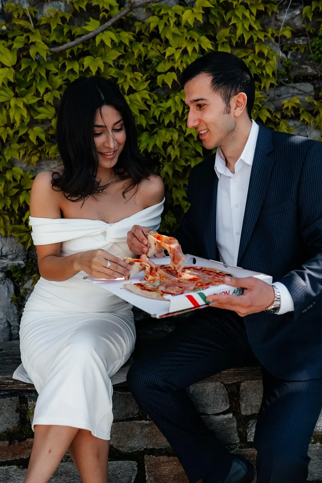 Omuzları açık beyaz elbiseli Zariyat ve lacivert takım elbiseli Ali, yeşil sarmaşığın önünde bir taş duvarda oturmuş, kutudan pizza paylaşıyor