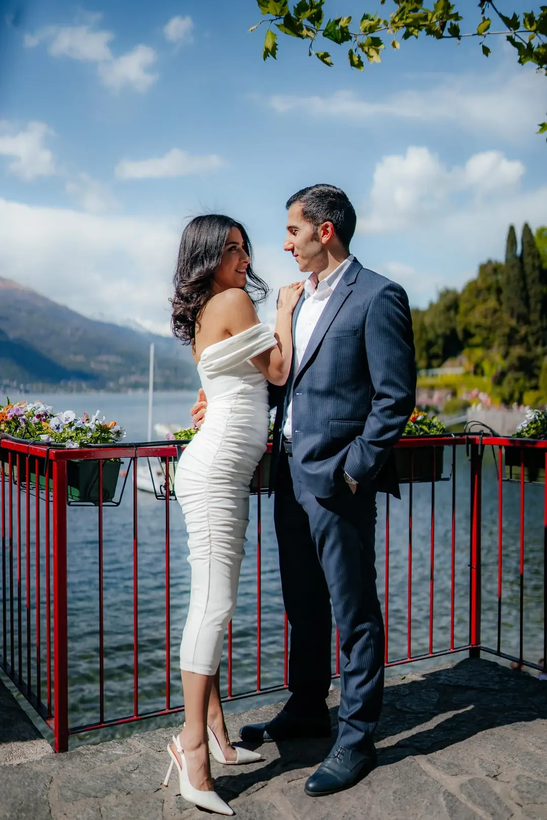 Ali e Zariyat su un balcone con la ringhiera rossa e le fioriere, il lago e le montagne alle spalle sotto un cielo azzurro