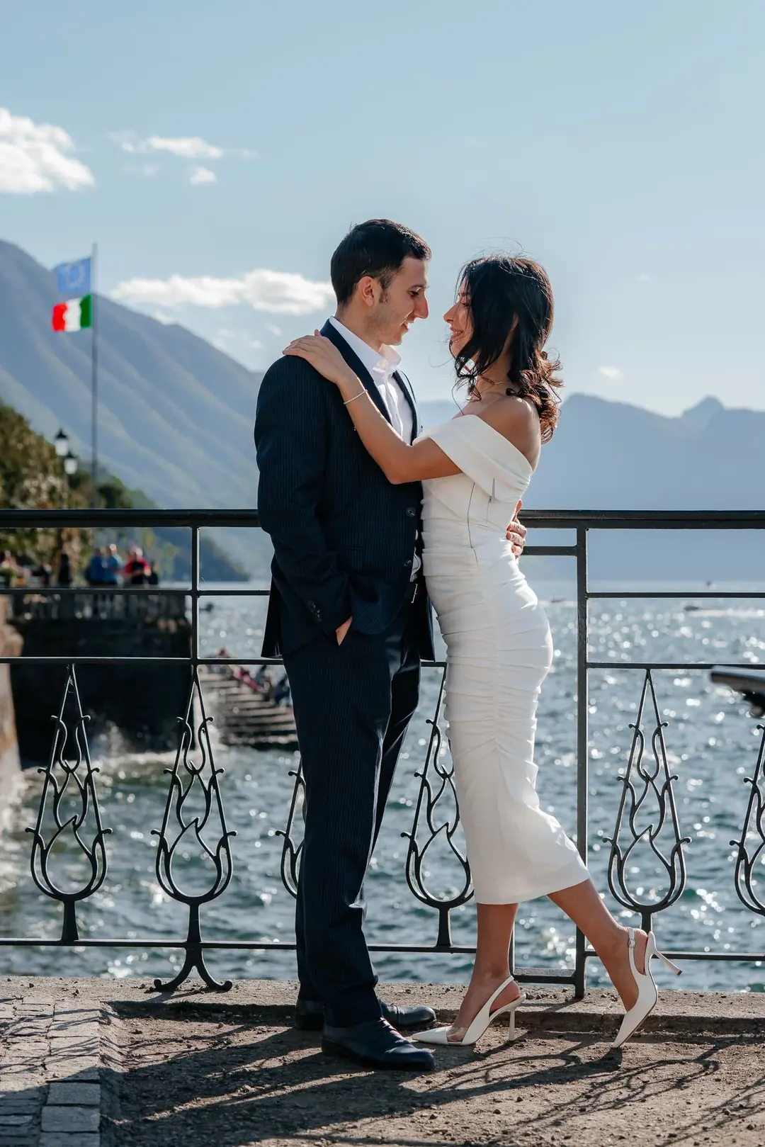 Ali and Zariyat embracing at a wrought-iron railing above Lake Como during their engagement session, mountains across the water