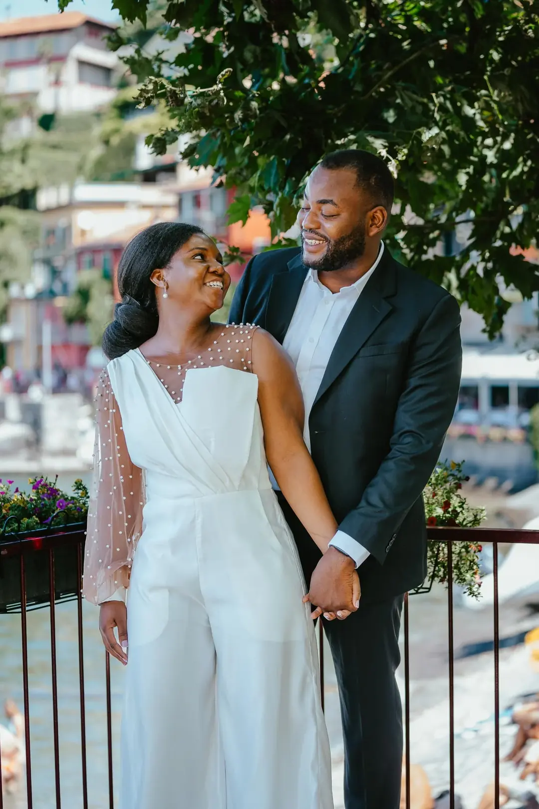 Daniel and Adekunbi smiling at each other at a railing with flower boxes, lakeside houses and boats blurred behind them