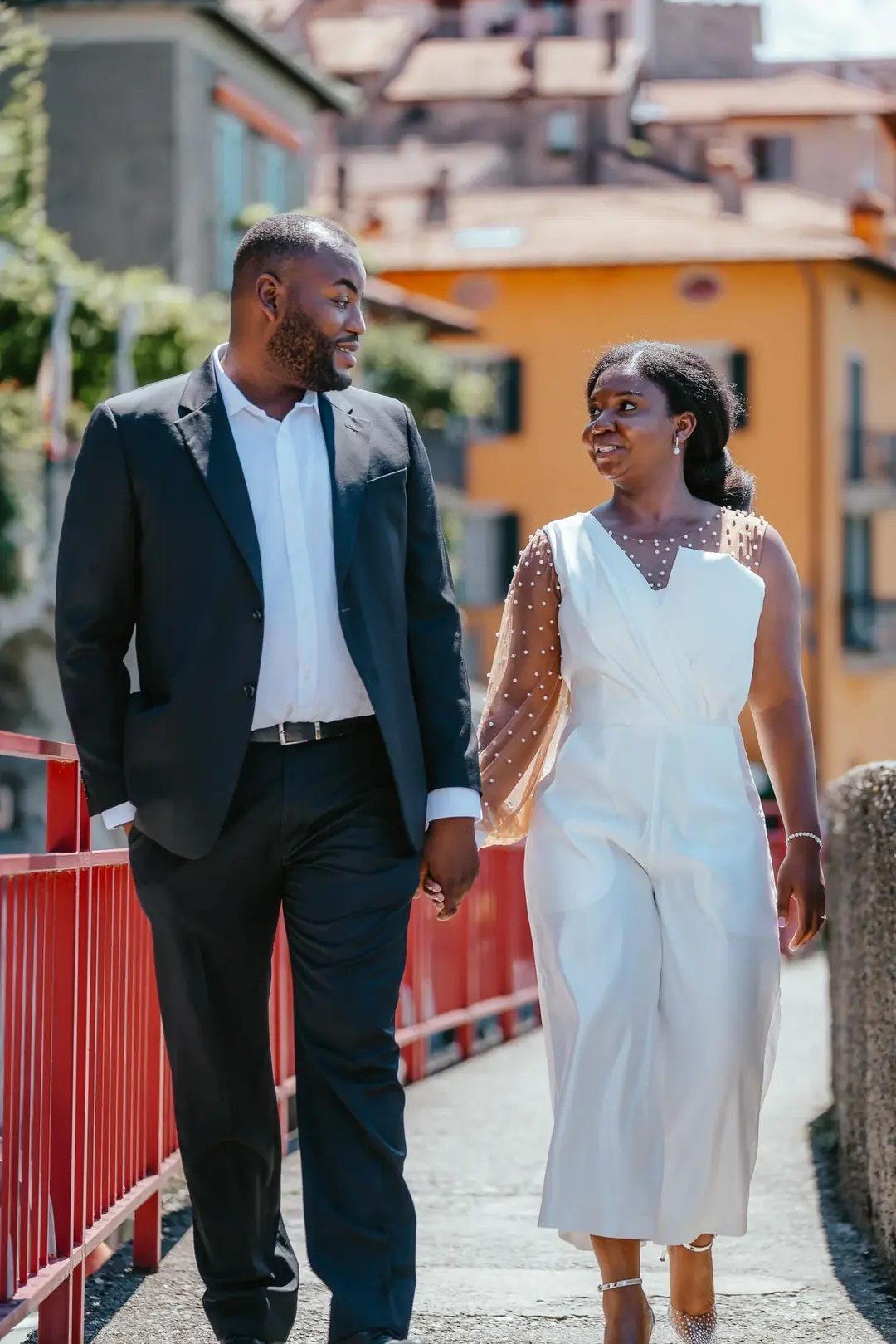 Daniel and Adekunbi walking hand in hand along a red railing in front of an orange house