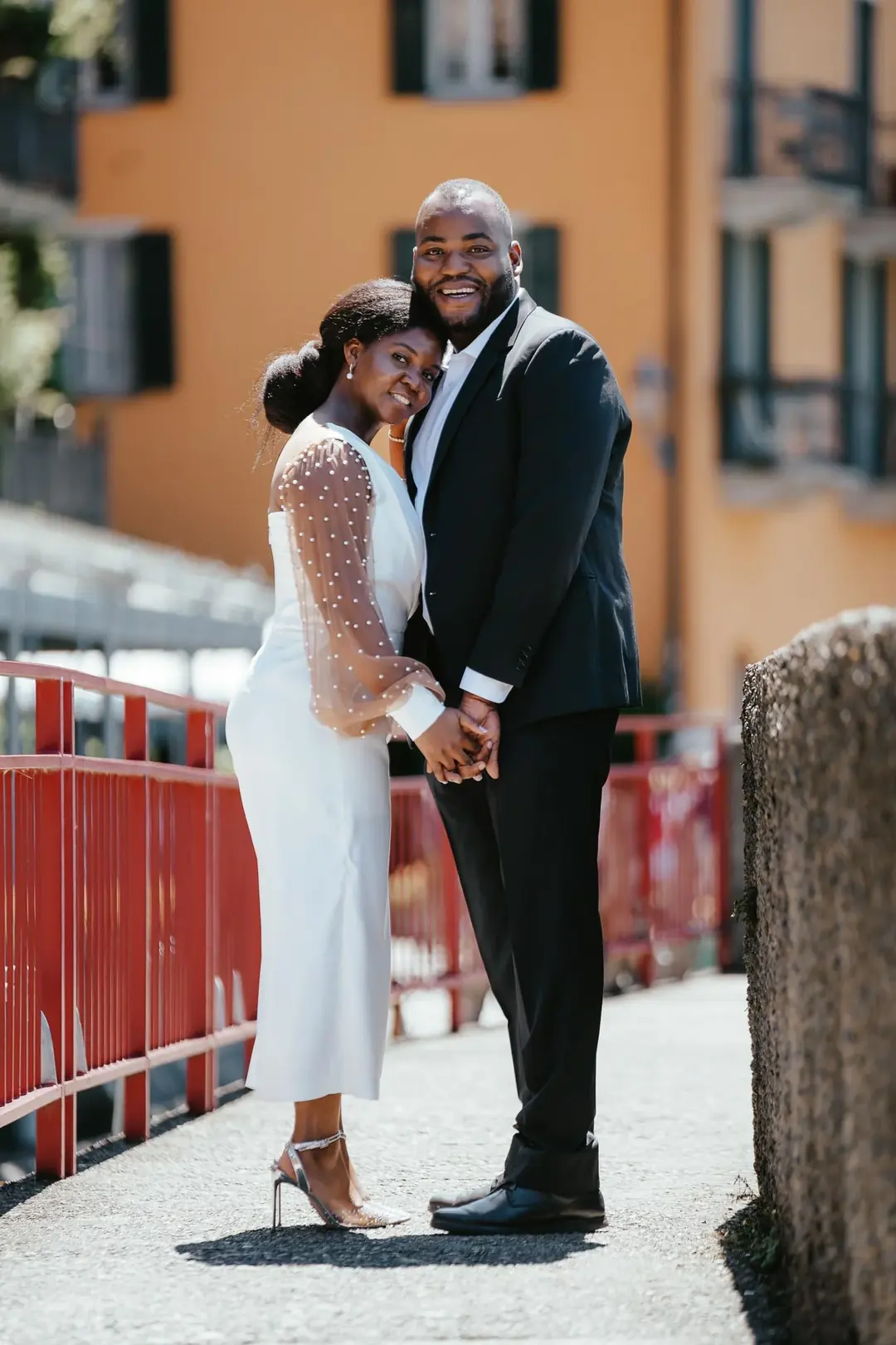 Daniel and Adekunbi standing close at a red railing in front of an orange house, holding hands