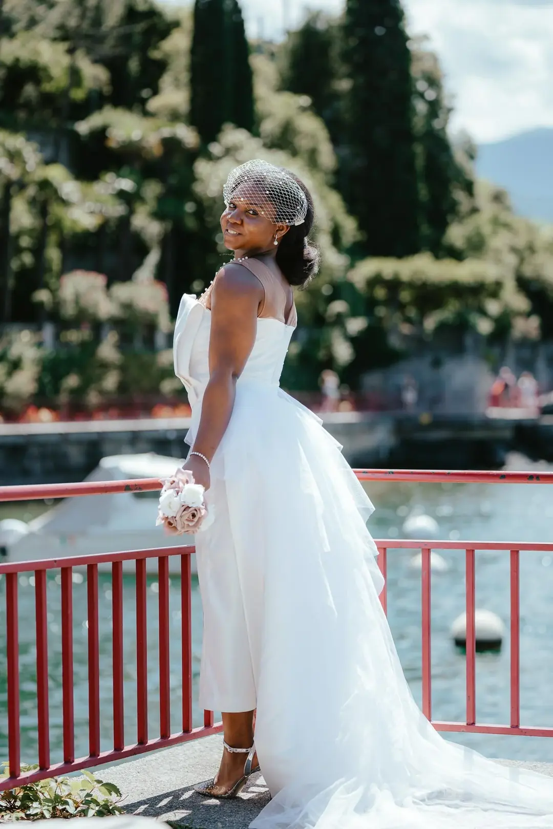 Adekunbi con una veletta si volta a guardare indietro a una ringhiera rossa sul lago, con in mano un bouquet rosa pallido