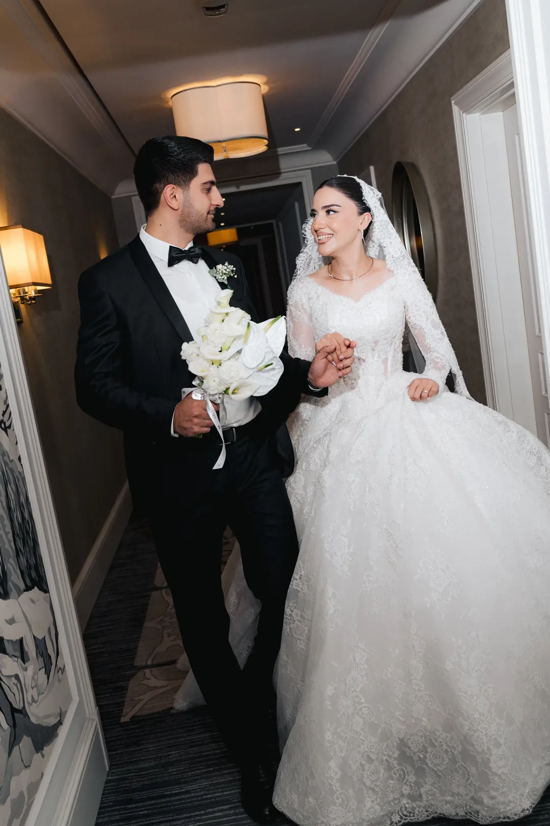 A bride and groom laughing as they walk hand in hand down a hotel corridor, he carrying her white bouquet