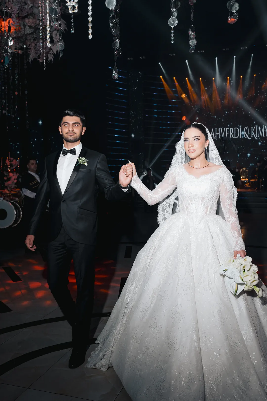 A bride and groom walking hand in hand onto a dance floor under stage lights, her lace gown and long veil trailing