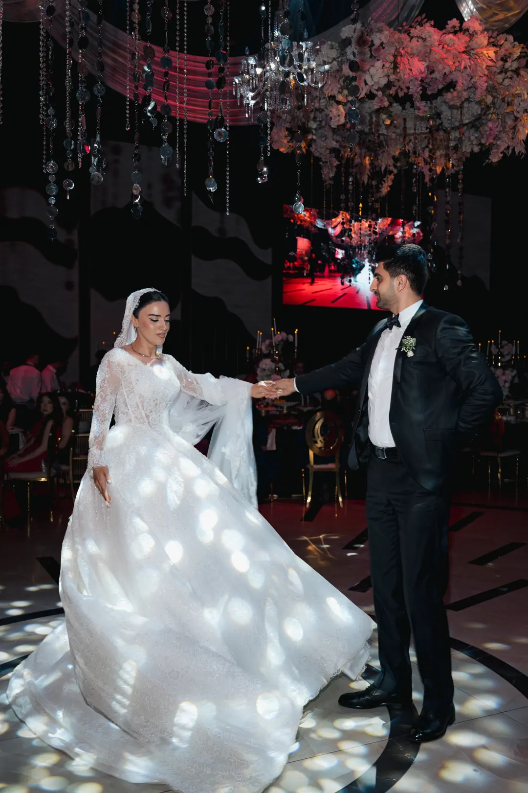 Allahverdi and Kimya dancing on a lit floor under hanging crystals, light spots across her full gown, guests behind them