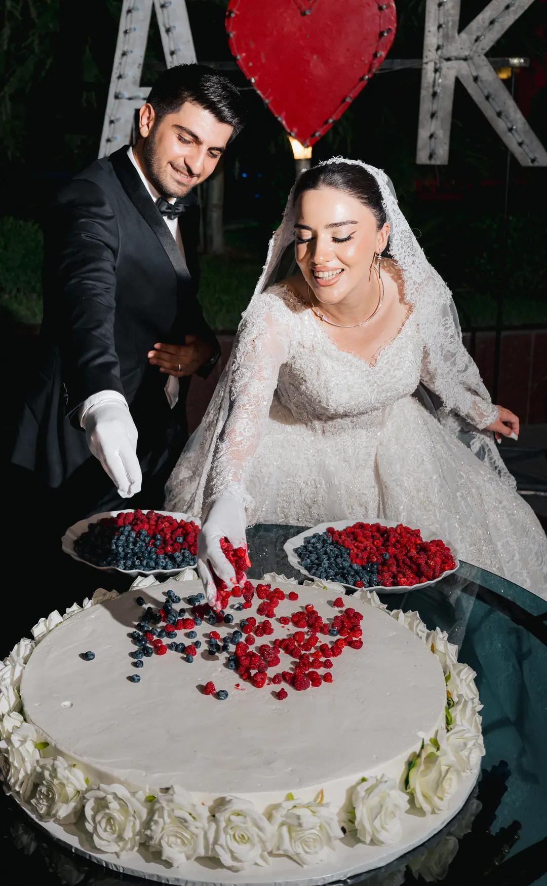 Kimya lascia cadere dei frutti di bosco su una torta nuziale bianca mentre Allahverdi allunga la mano accanto a lei, entrambi con guanti bianchi