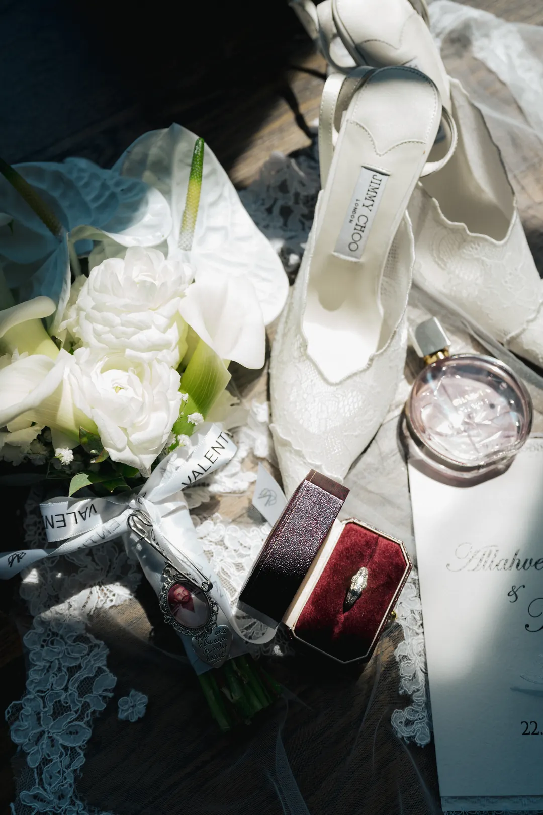 A flat lay of a bride's lace shoes, white flowers, a ring in a red box, a perfume bottle and a printed invitation in hard sunlight