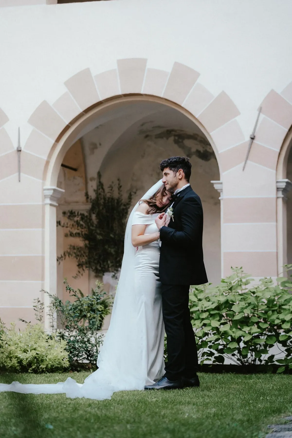 Musa and Solmaz embracing beneath an arched loggia in Susa, Piedmont, her long veil trailing across the lawn, green shrubs around them