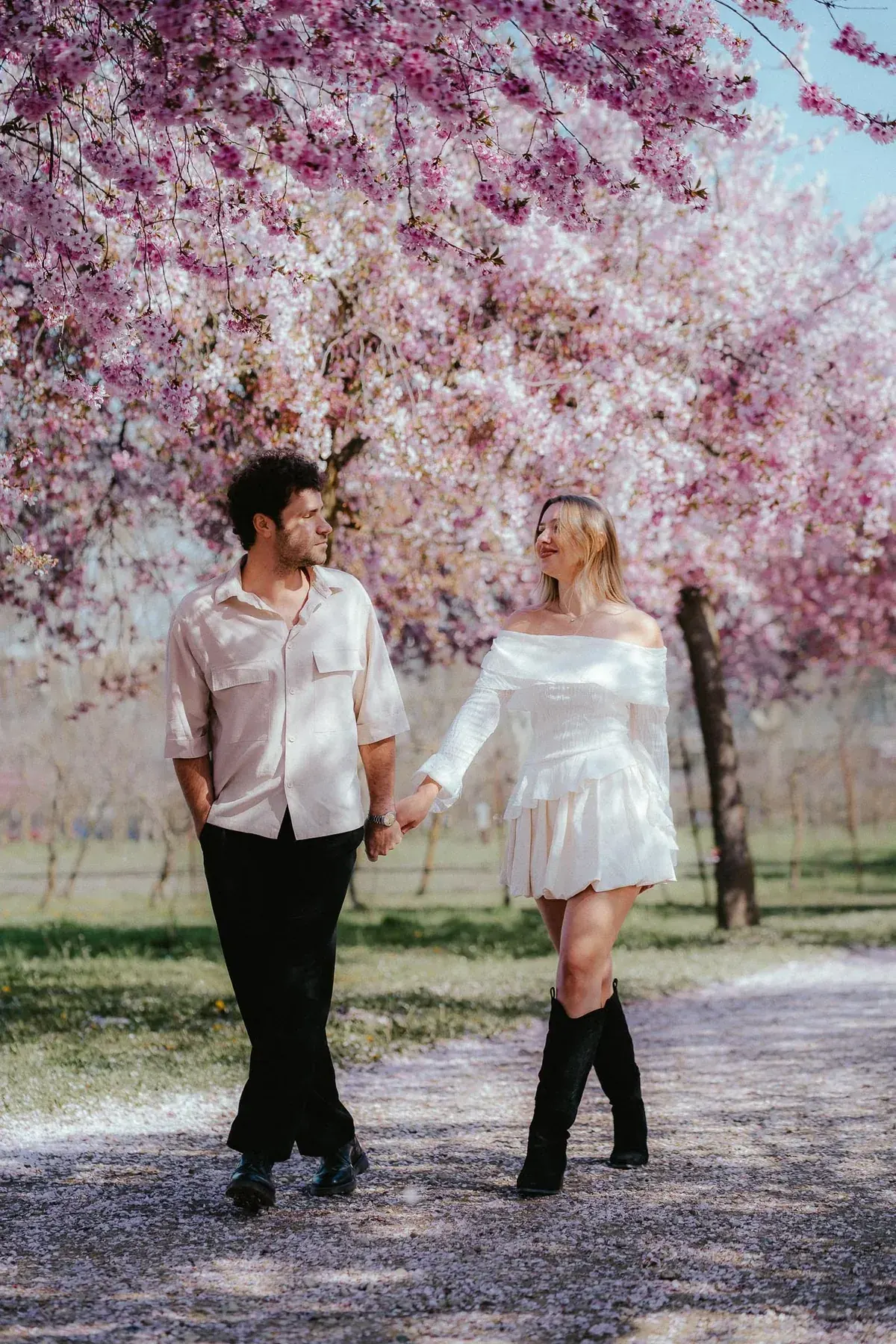 Gabriele and Maddalena walking hand in hand under pink cherry blossom in a Turin park, petals on the path