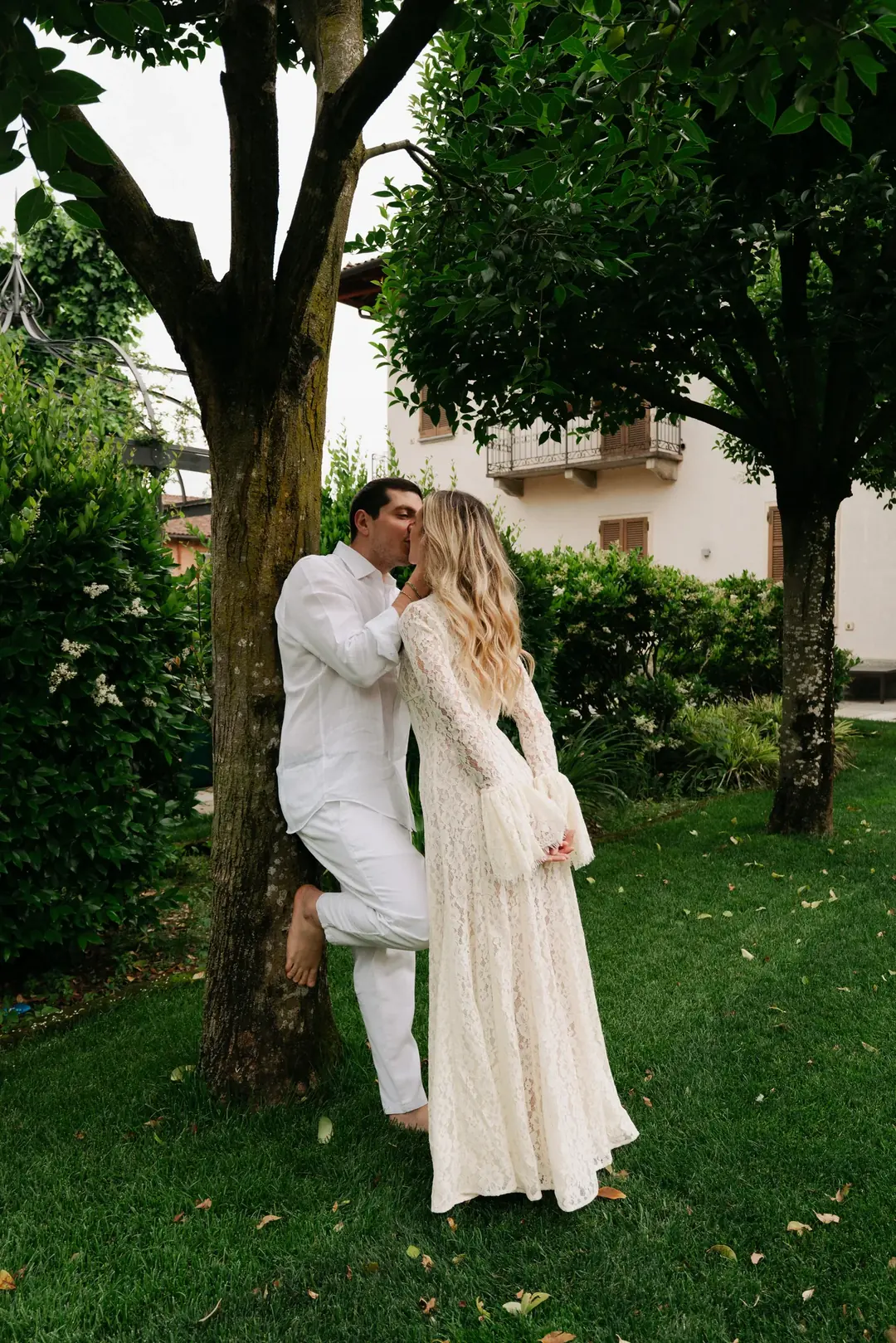Andrea kissing Daria as he leans barefoot against a tree in a green garden in Turin, she in a long lace dress