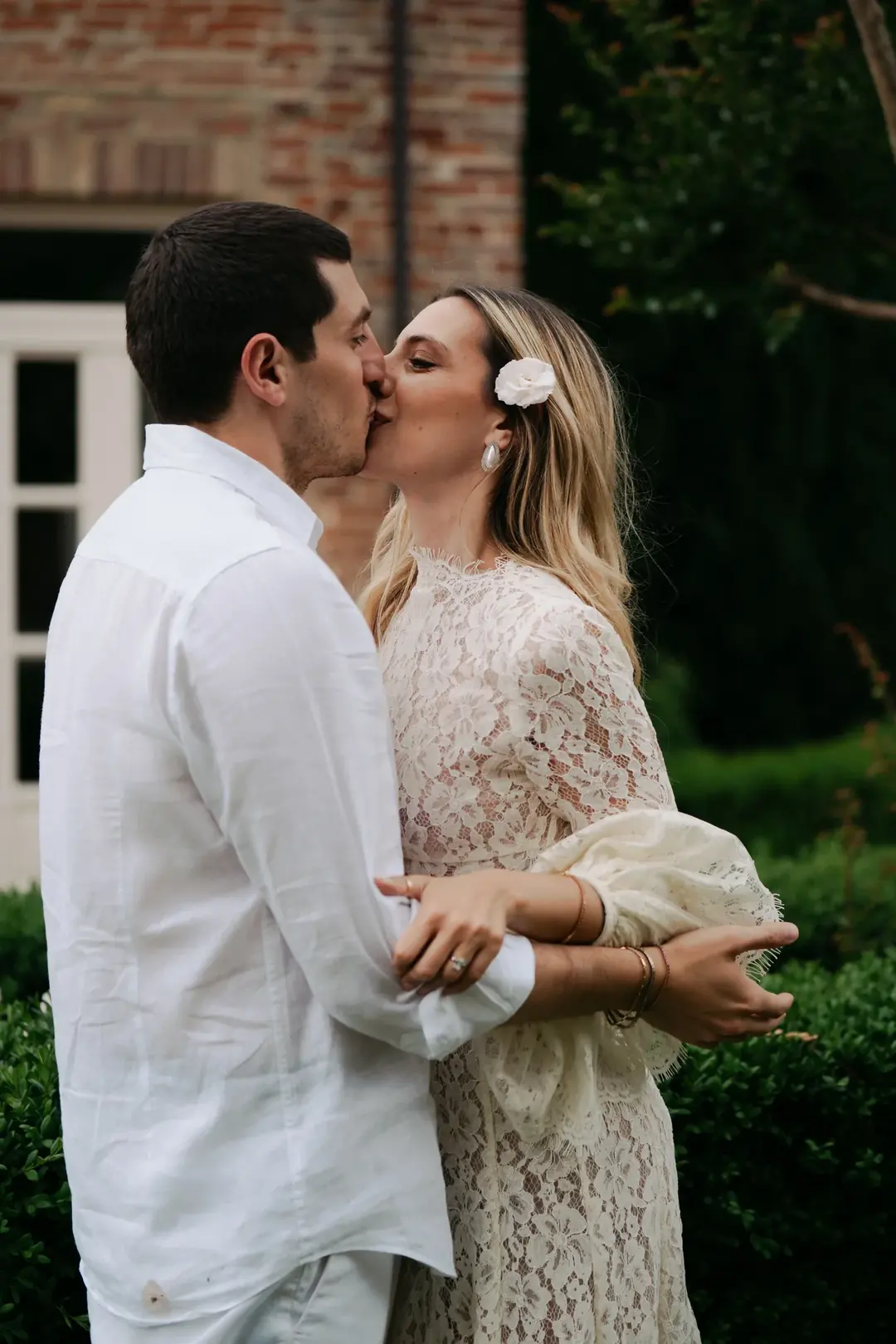 Daria and Andrea kissing in a garden beside a brick house, arms linked, she in a lace dress