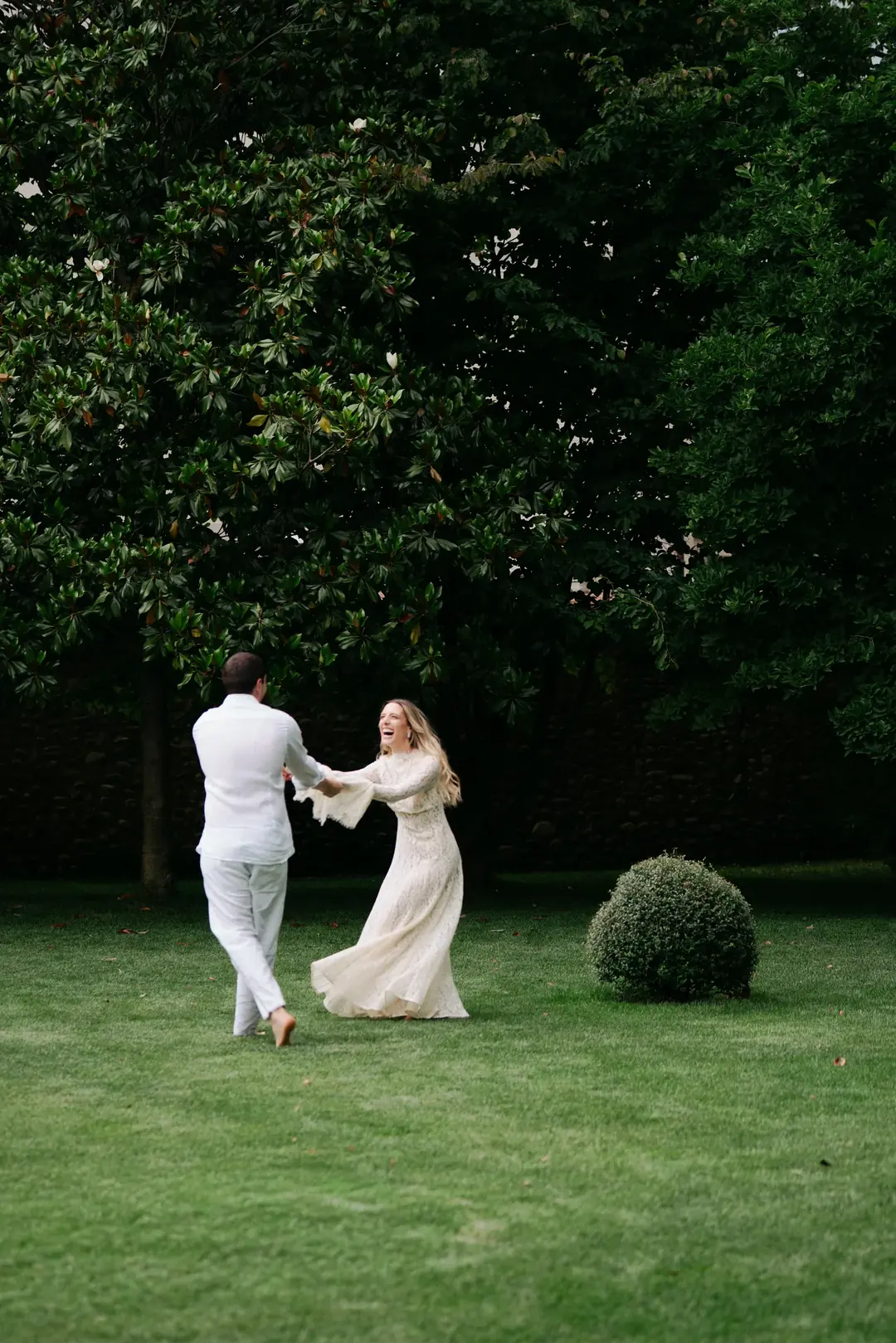 Daria and Andrea holding hands and swinging each other round on a wide green lawn, a large tree and a round clipped bush beside them