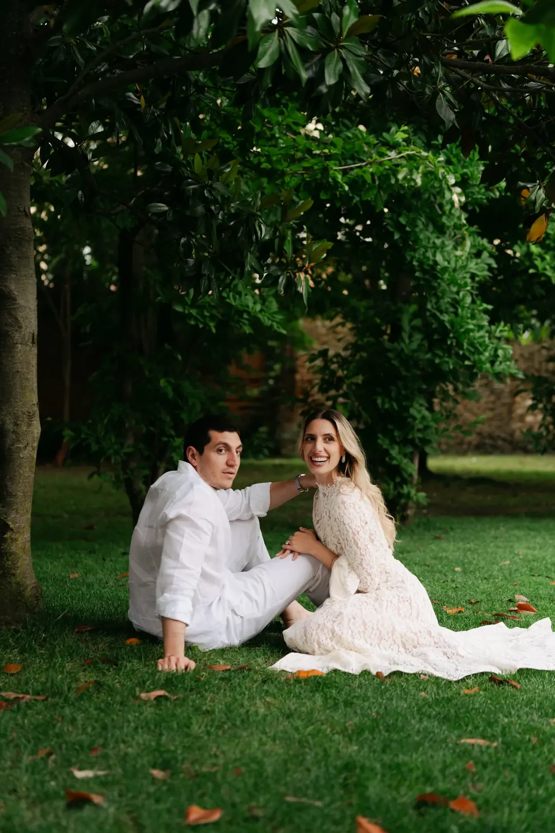 Daria laughing towards the camera as she and Andrea sit on a lawn under trees, her lace dress spread on the grass