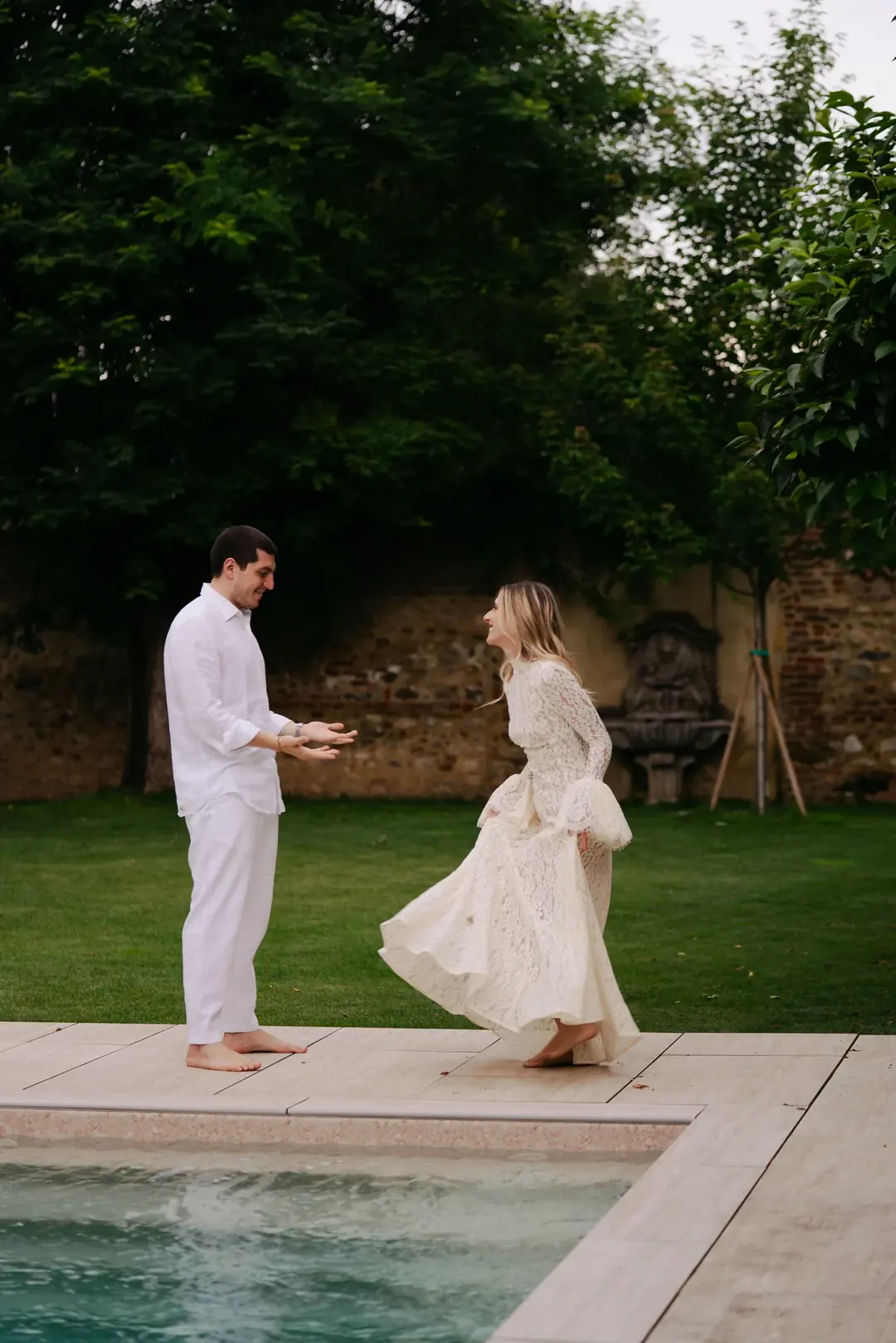 Daria and Andrea barefoot at the edge of a garden pool, she turning in a long lace dress as he reaches for her, an old brick wall behind them