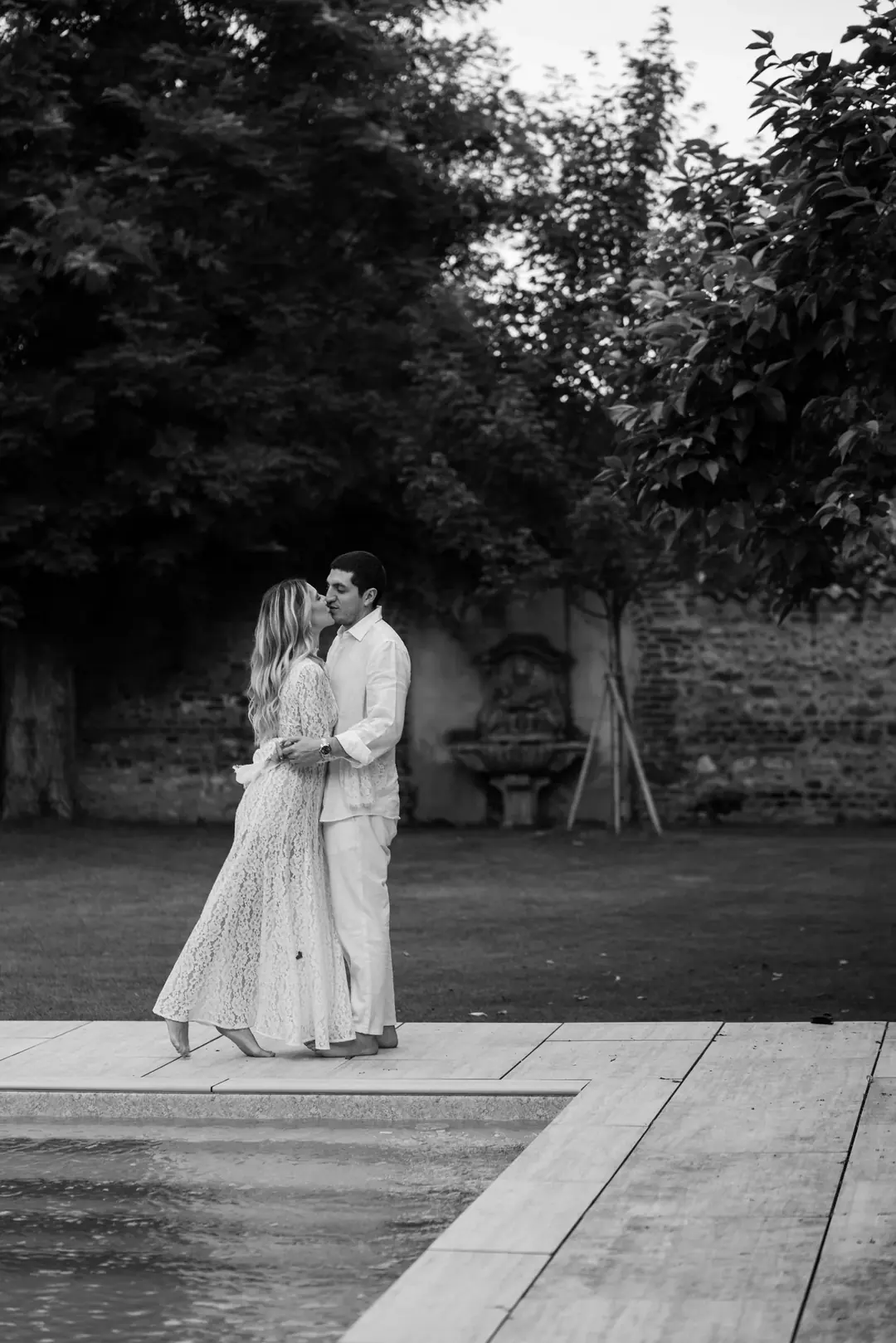 Daria and Andrea kissing by a garden pool, an old brick wall with a stone fountain behind them, in black and white
