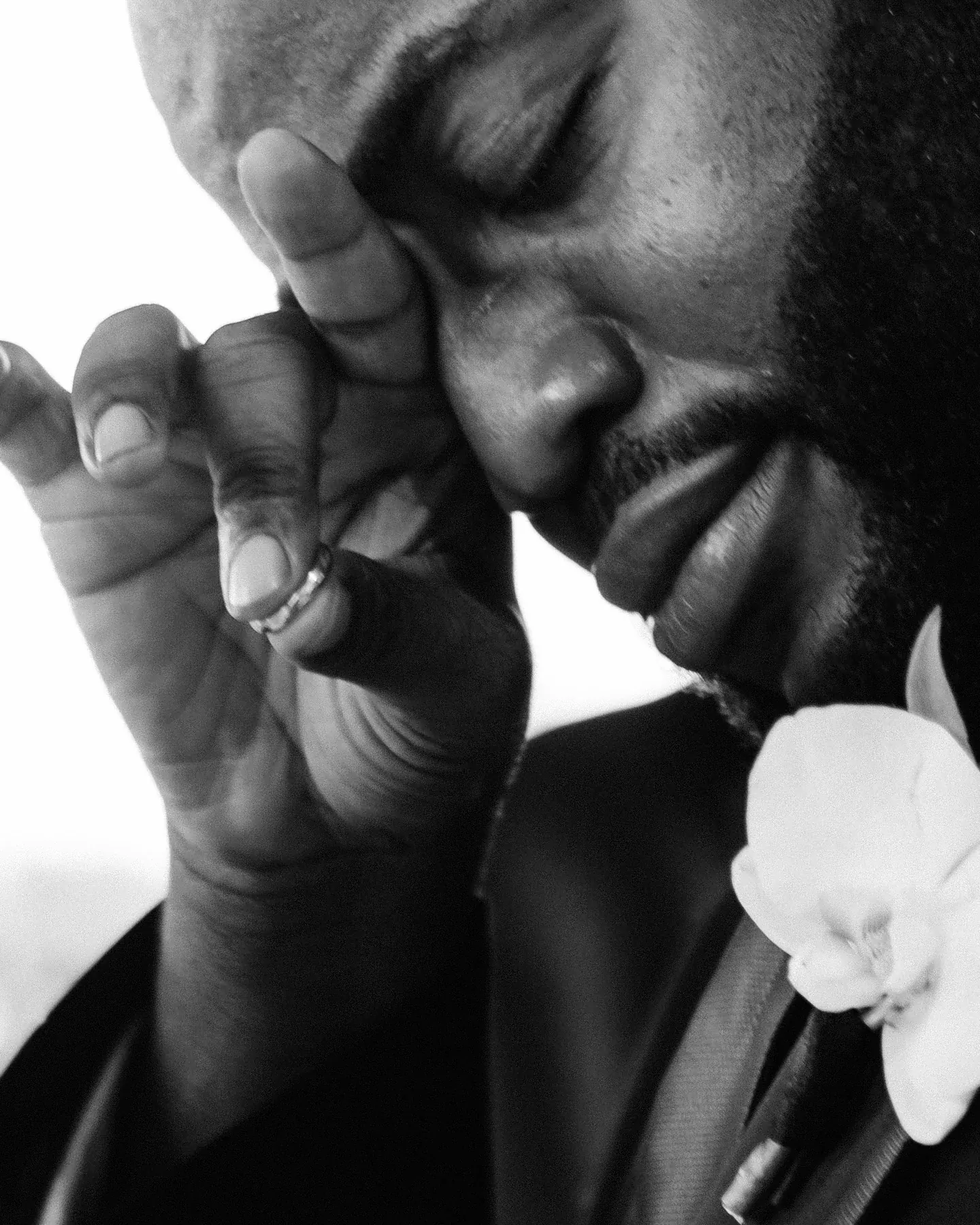 A man in a dark suit with a white orchid on his lapel presses his fingers to his closed eye, in black and white