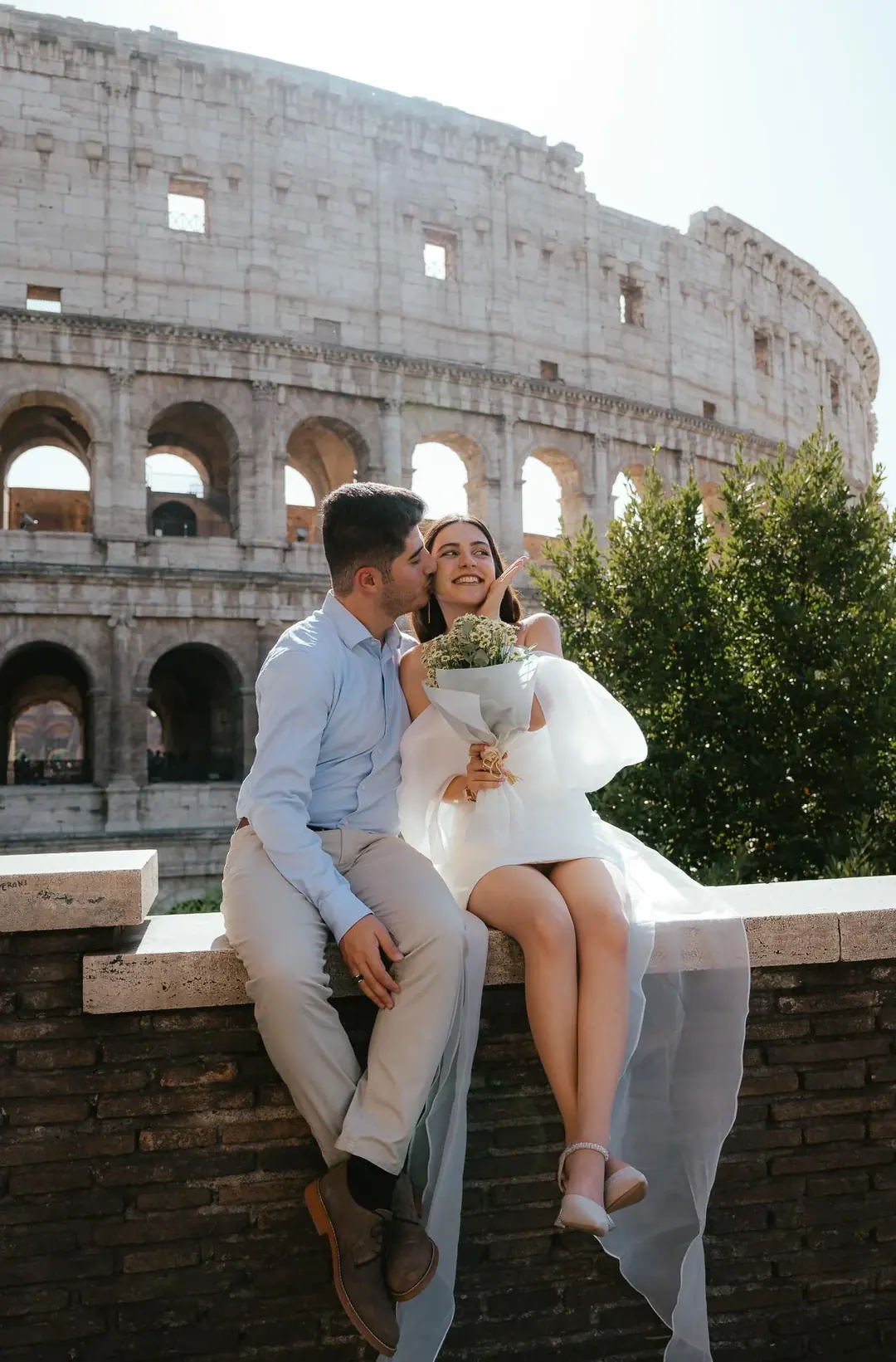 A couple sitting on a stone parapet facing the Colosseum in Rome, he kisses her cheek as she laughs, holding a bouquet of daisies