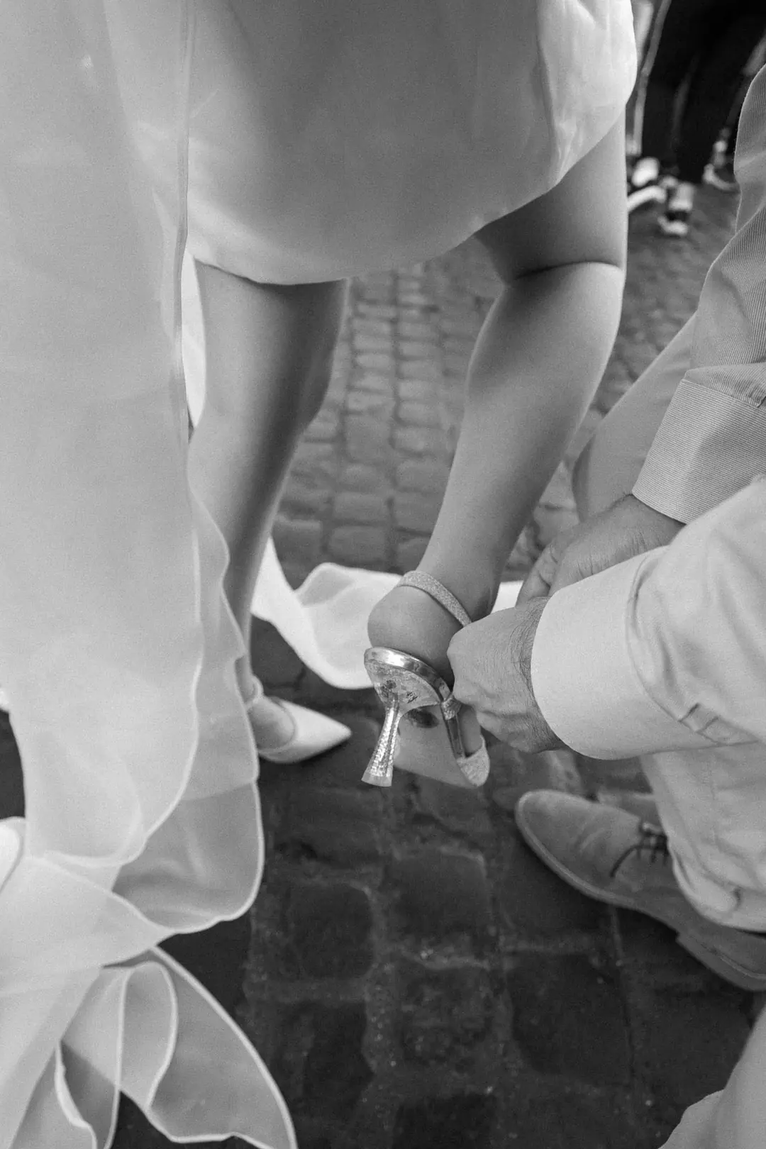 A man kneeling on a cobbled street to fasten a woman's high-heeled shoe, her organza skirt in the foreground, black and white