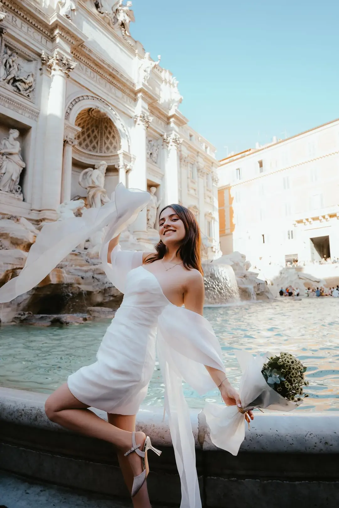 A woman in a short white dress with sheer organza sleeves flying, one heel raised and a bouquet in her hand, smiling in front of the Trevi Fountain in Rome, other visitors at the basin behind