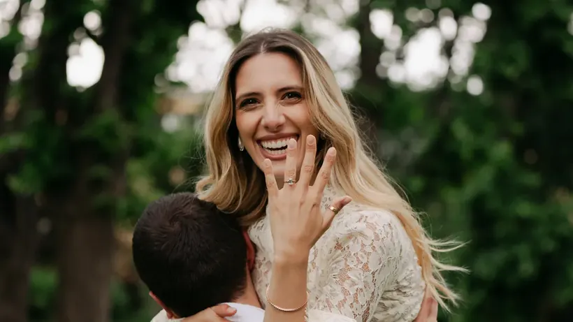 Daria laughing as she holds up her hand to show her engagement ring, while Andrea lifts her in a green garden