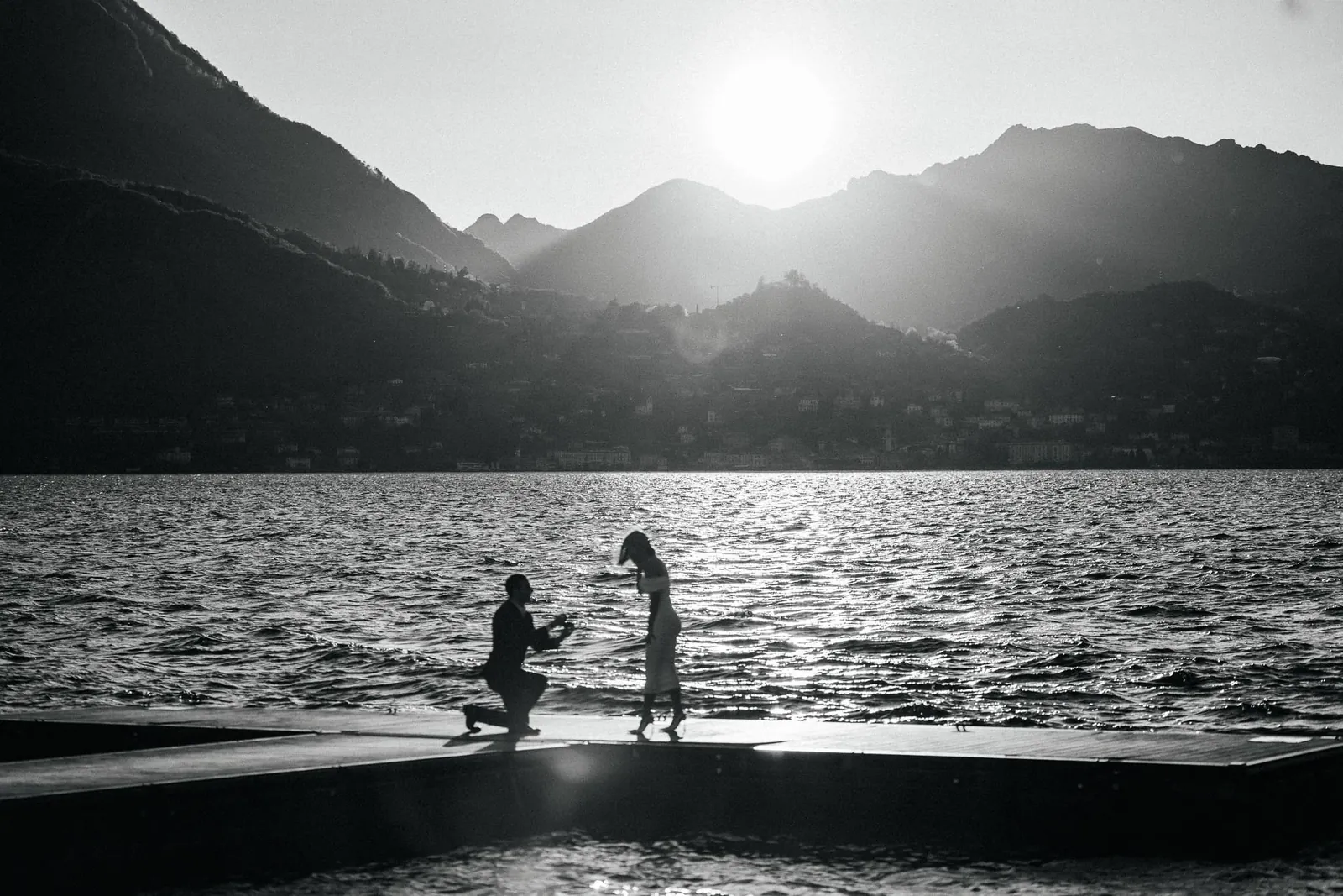 Un uomo inginocchiato davanti a una donna su un pontile, entrambi in controluce contro il sole basso sopra le montagne e il lago