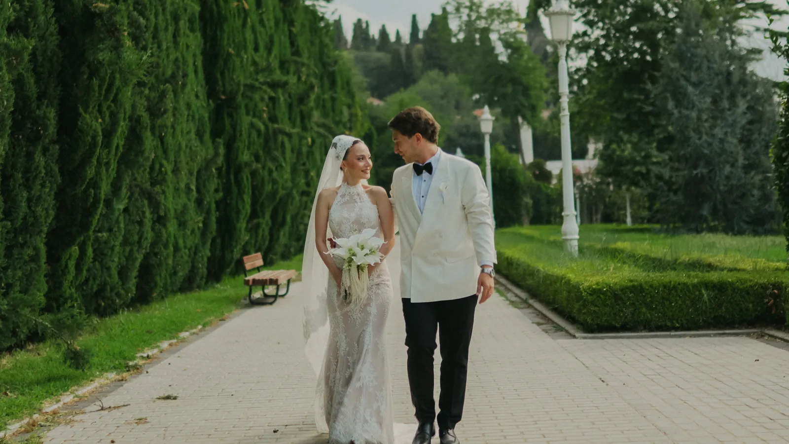 Gül and Oğuz walking along a paved garden path between a tall cypress hedge and clipped box borders, she holding white calla lilies