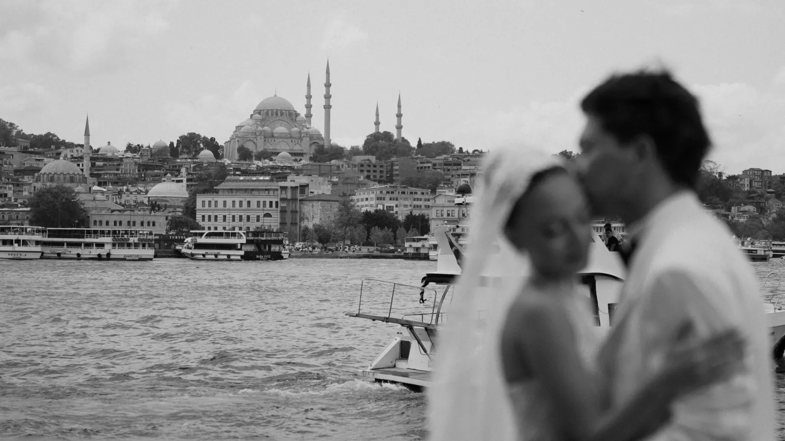 Black and white frame of Gül and Oğuz embracing in soft focus in the foreground, with ferries and a domed mosque with four minarets across the water behind them