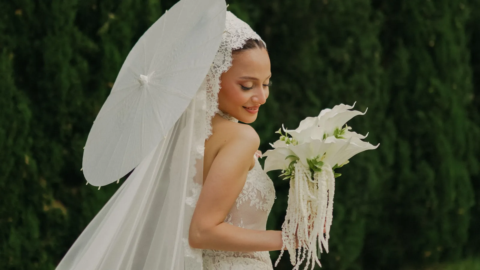 Gül sorride guardando un bouquet di calle bianche sotto un ombrellino di carta bianco, con il velo di pizzo che le scende oltre la spalla