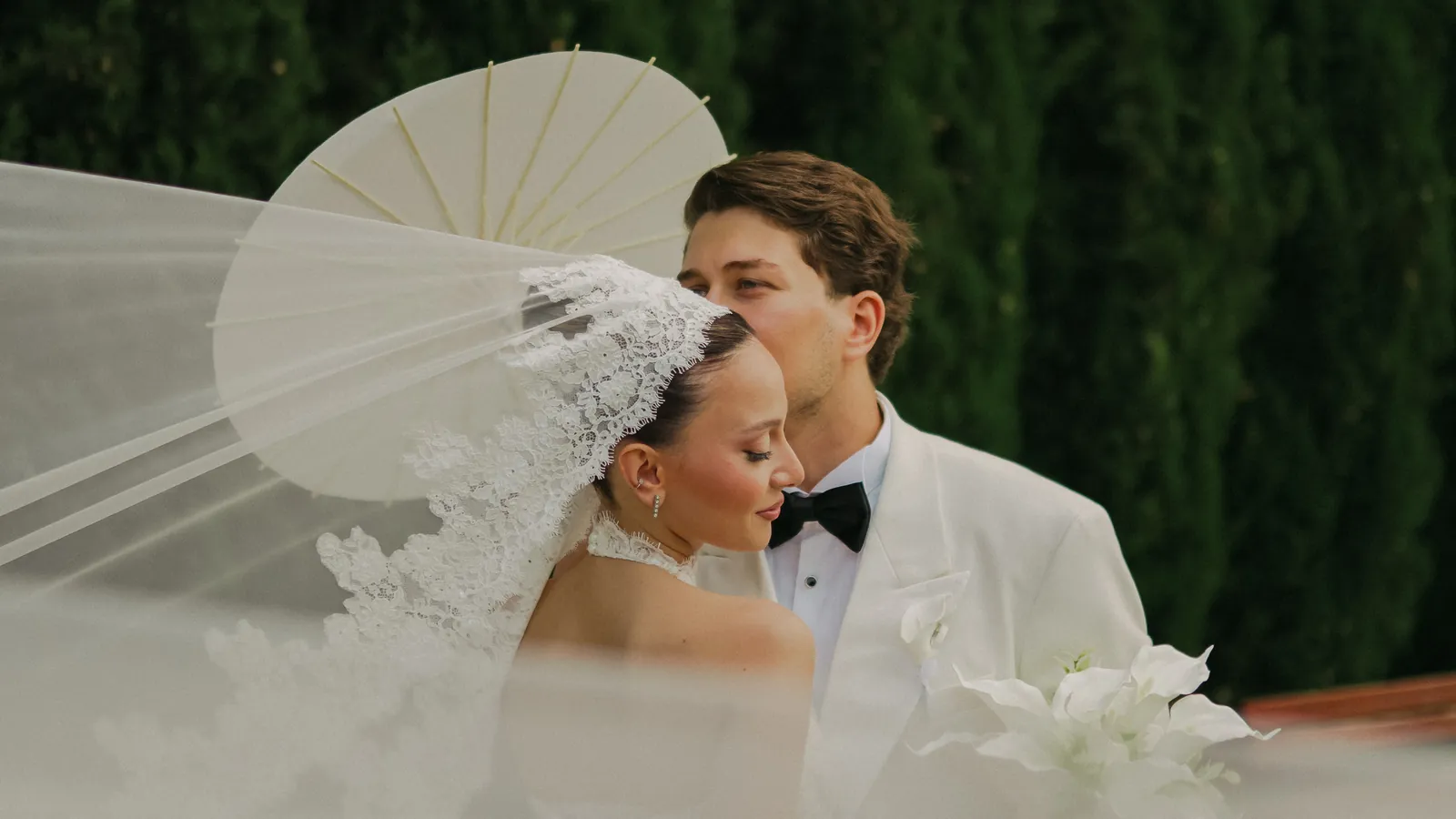 Gül holding a white parasol as her lace veil lifts in the wind, Oğuz in a white dinner jacket leaning in behind her, a cypress hedge behind them