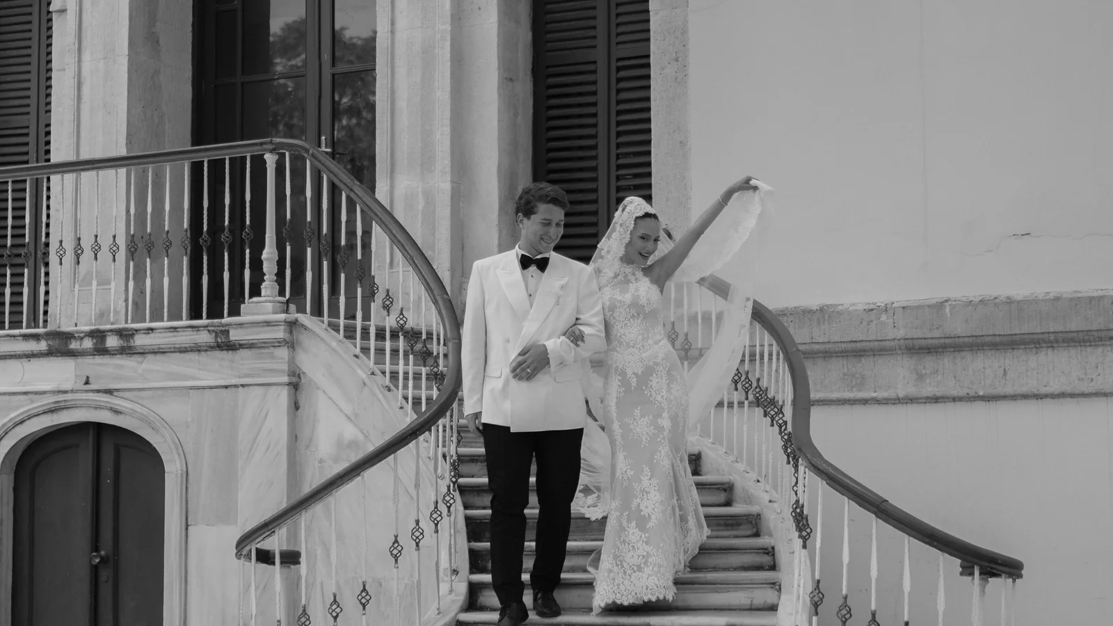 Gül and Oğuz walking down a curved marble staircase together, Gül lifting her veil high with one hand, black and white
