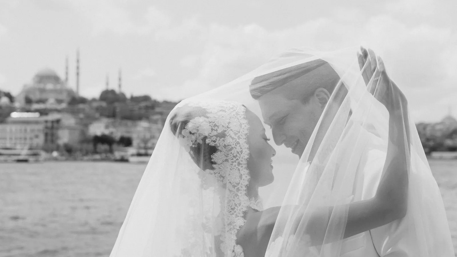 Gül e Oğuz viso a viso sotto il velo di pizzo di lei, in riva all'acqua, con una moschea a cupola sull'altra sponda, in bianco e nero