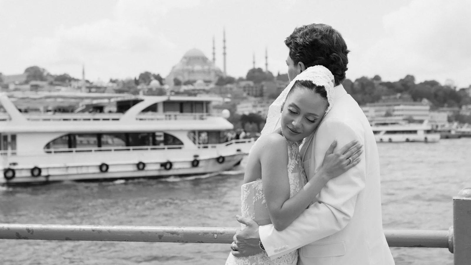 Gül and Oğuz embracing at a railing by the water in Istanbul, a ferry passing and domes and minarets on the far shore, black and white